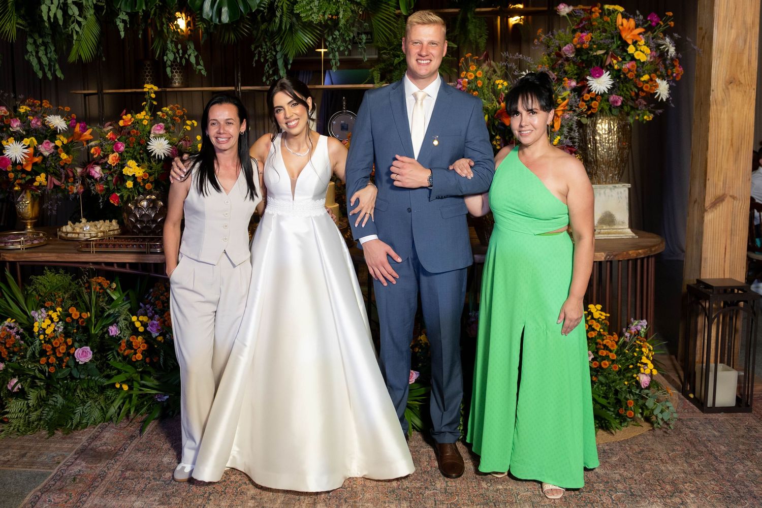 Bride in white gown poses with groom in suit and two women in green and gray at floral wedding reception.