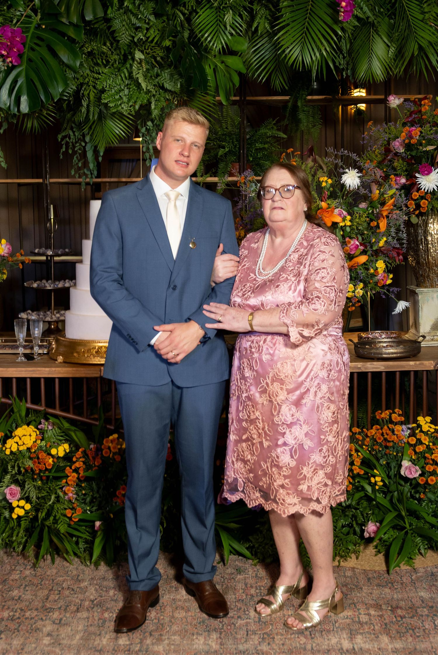 Man in blue suit and woman in pink lace dress pose together at a floral event backdrop.