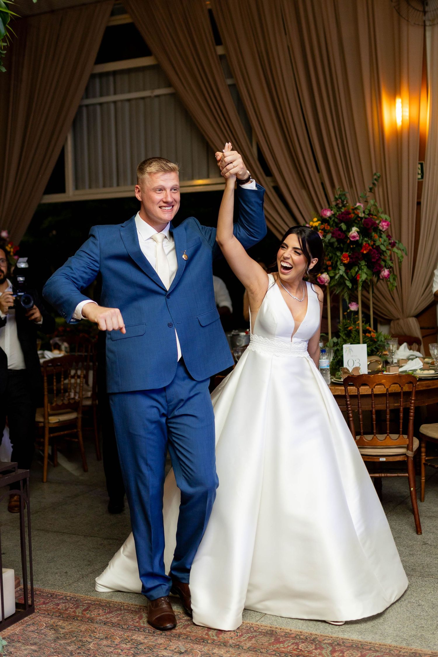 Joyful bride in white gown and groom in blue suit raising hands together at wedding reception entrance.