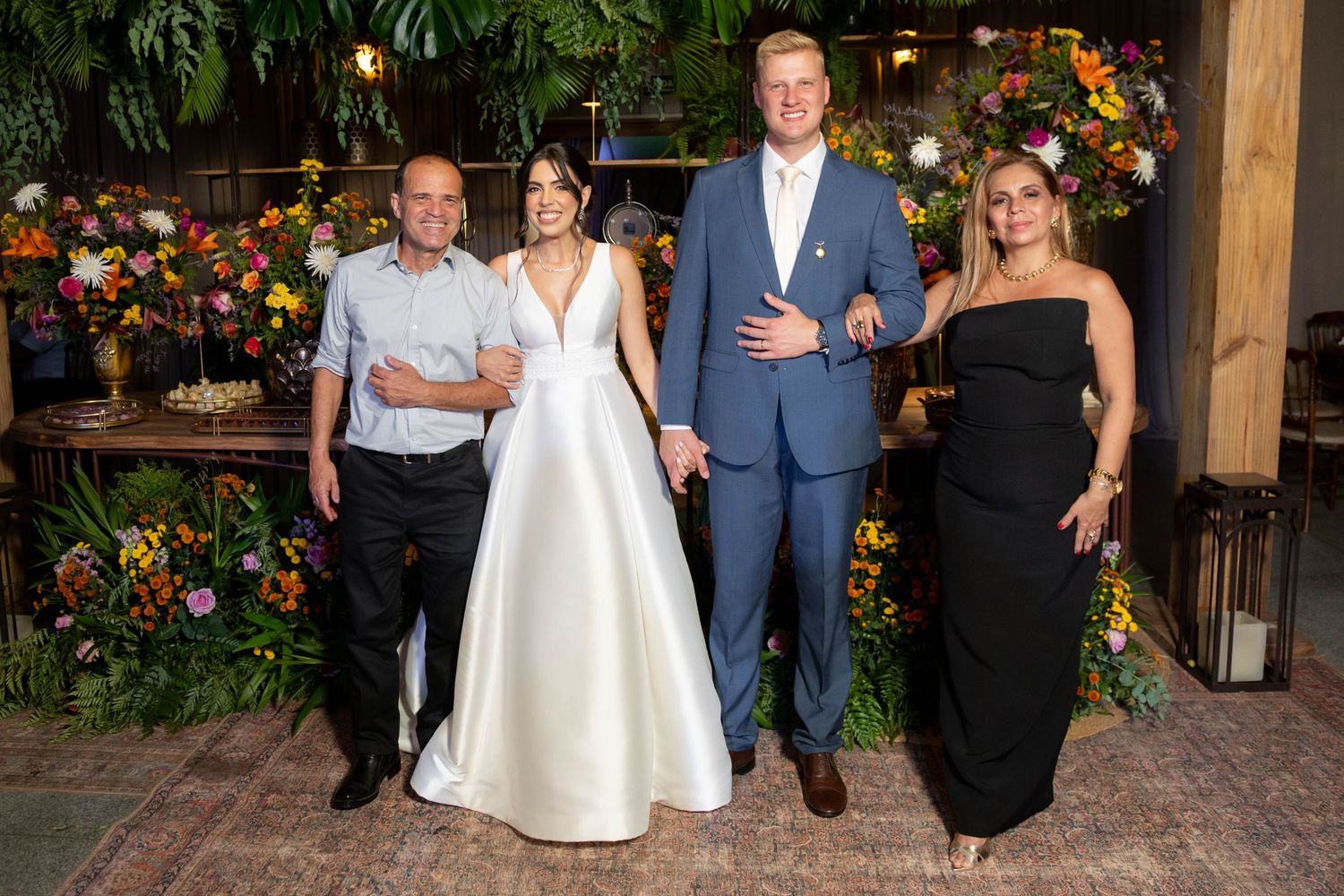 Bride in white gown and groom in blue suit pose with two guests amid lush floral decor at elegant wedding reception.