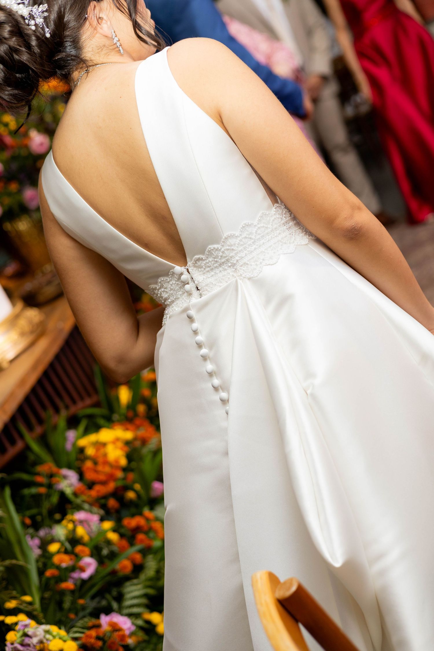 Bride in elegant white backless wedding dress with lace detail and pearl buttons, standing among colorful flowers.