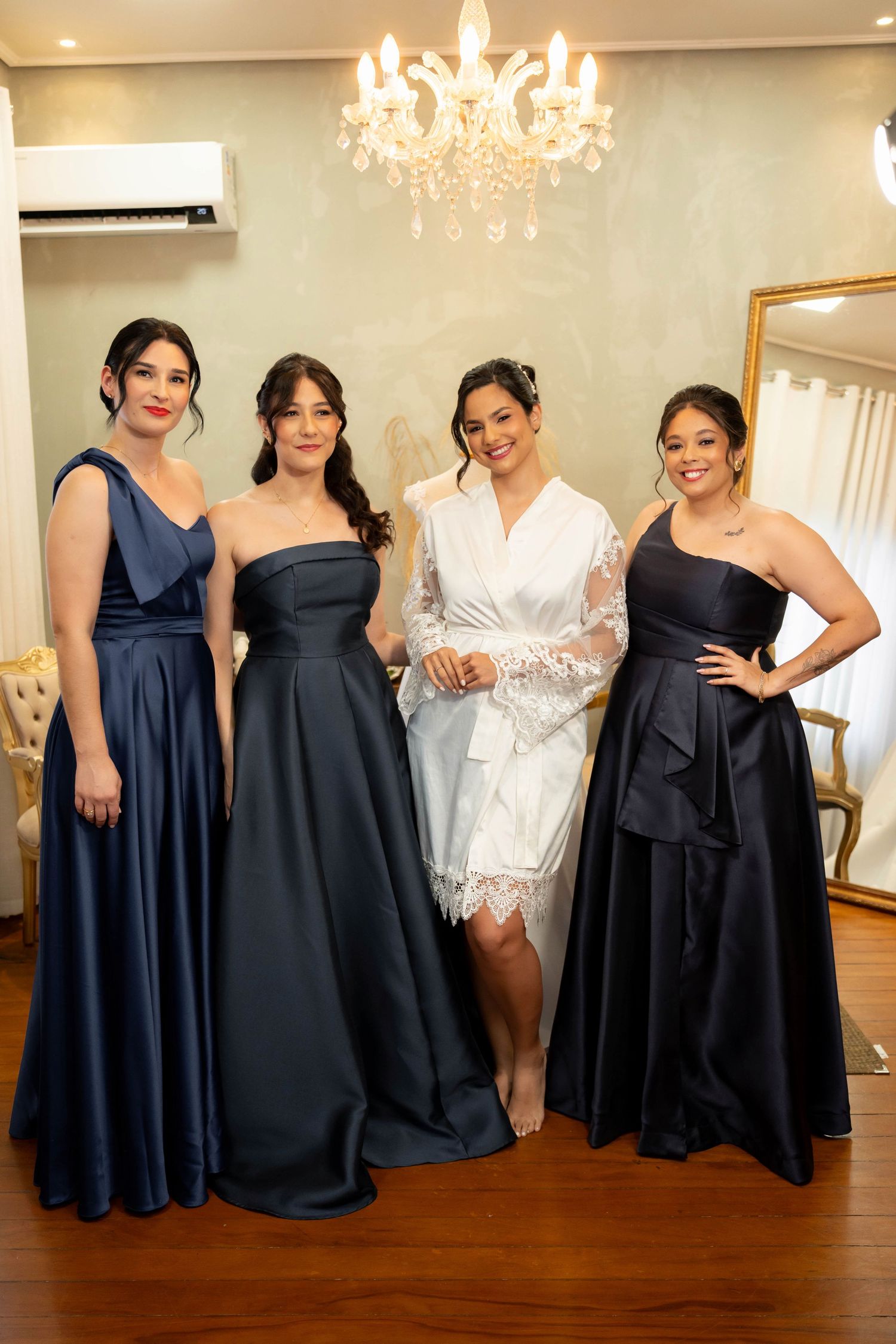 Bride in white robe poses with three bridesmaids in navy blue gowns inside an elegant dressing room.