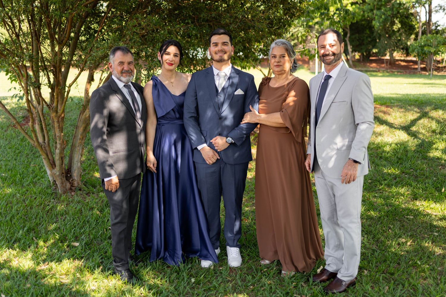 Family of five posing outdoors in formal attire, smiling together under lush green trees on a sunny day.