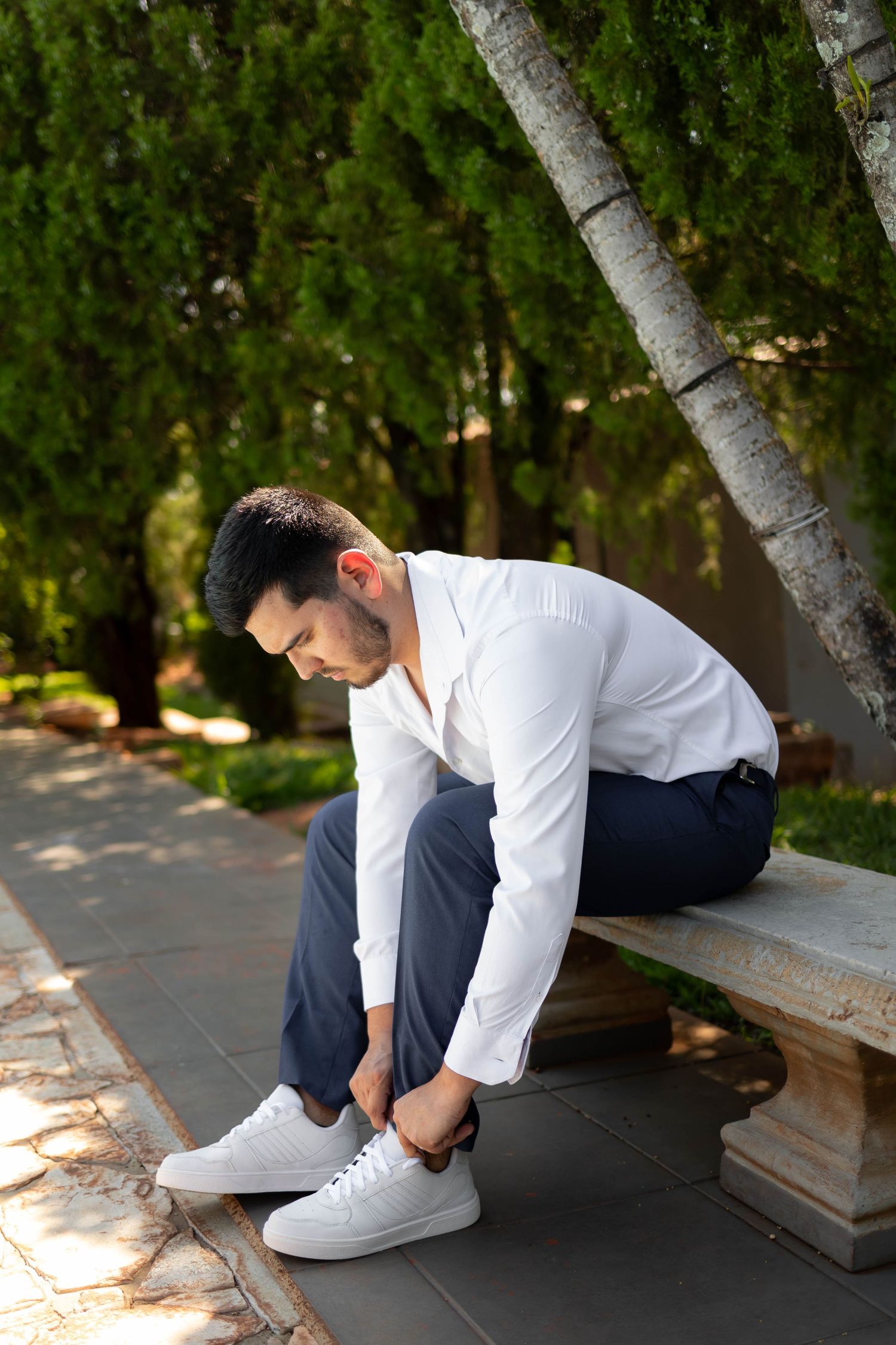 Man in white shirt and navy pants tying white sneakers on stone bench in lush green park.