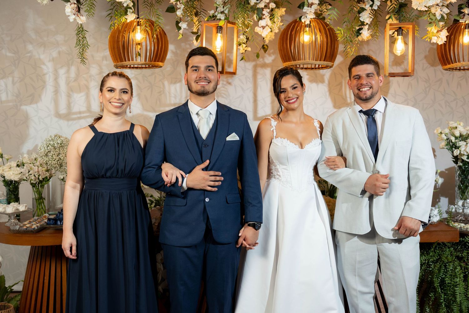 Two couples pose elegantly at a wedding, bride in white gown, groom in navy suit, flanked by guests in formal attire.