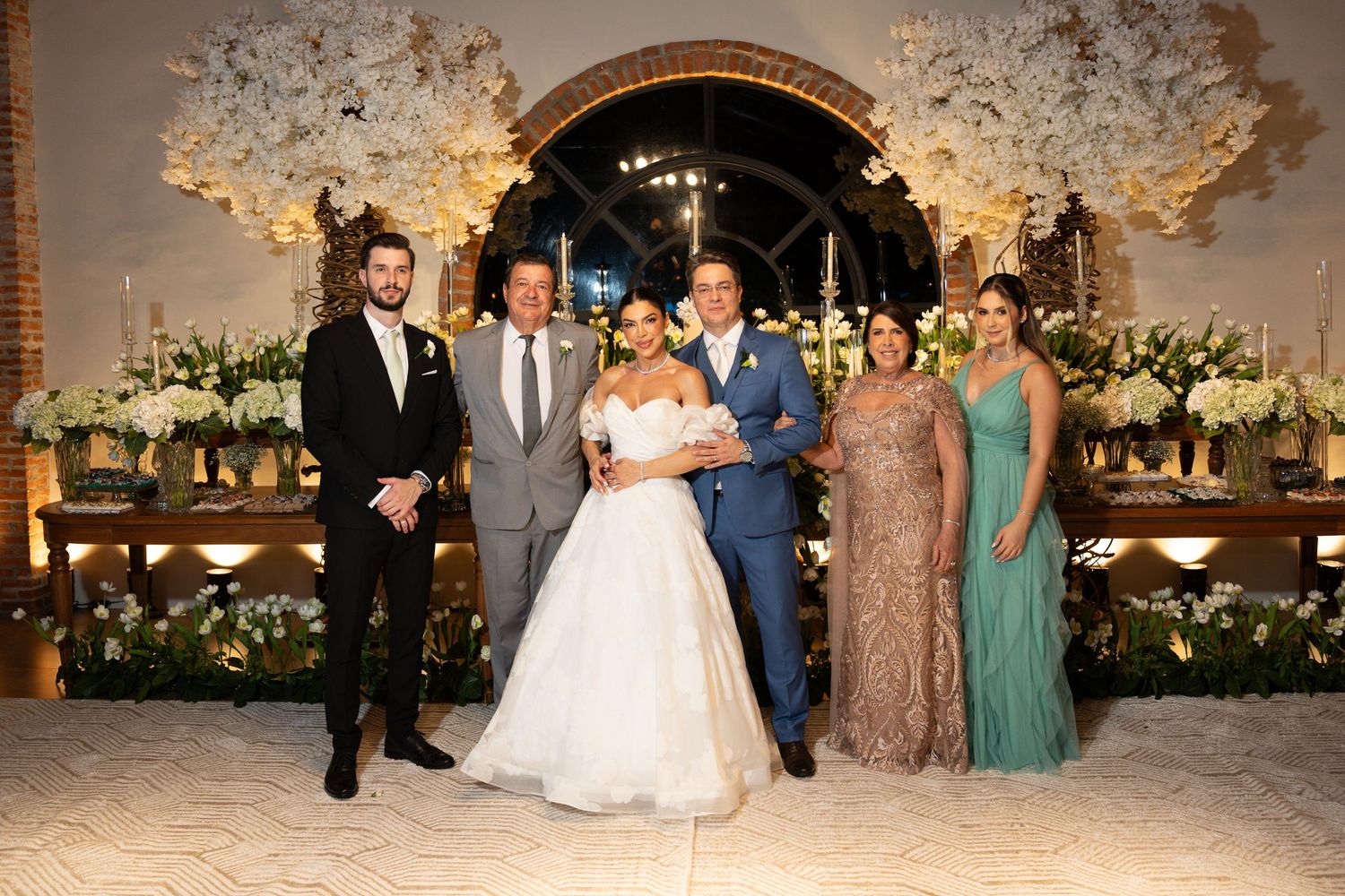 Wedding party poses before floral arch with bride in white gown, groom in blue suit, and elegantly dressed family members.