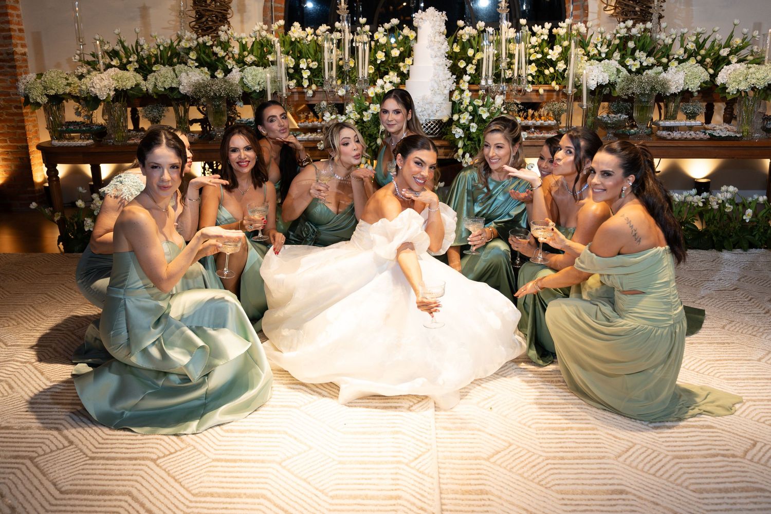 Bride in white gown poses with bridesmaids in sage green dresses on dance floor at elegant floral wedding reception.