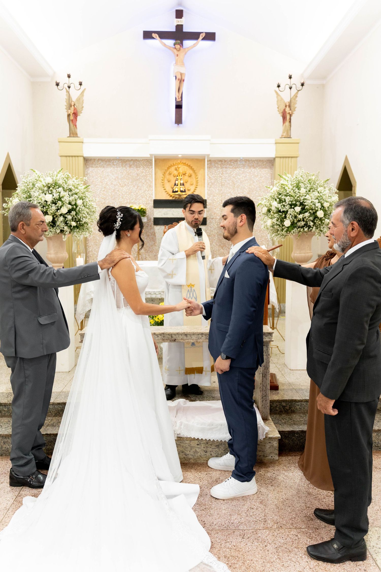 Bride and groom exchange vows at a Catholic church altar, flanked by witnesses, with a crucifix and floral arrangements behind.