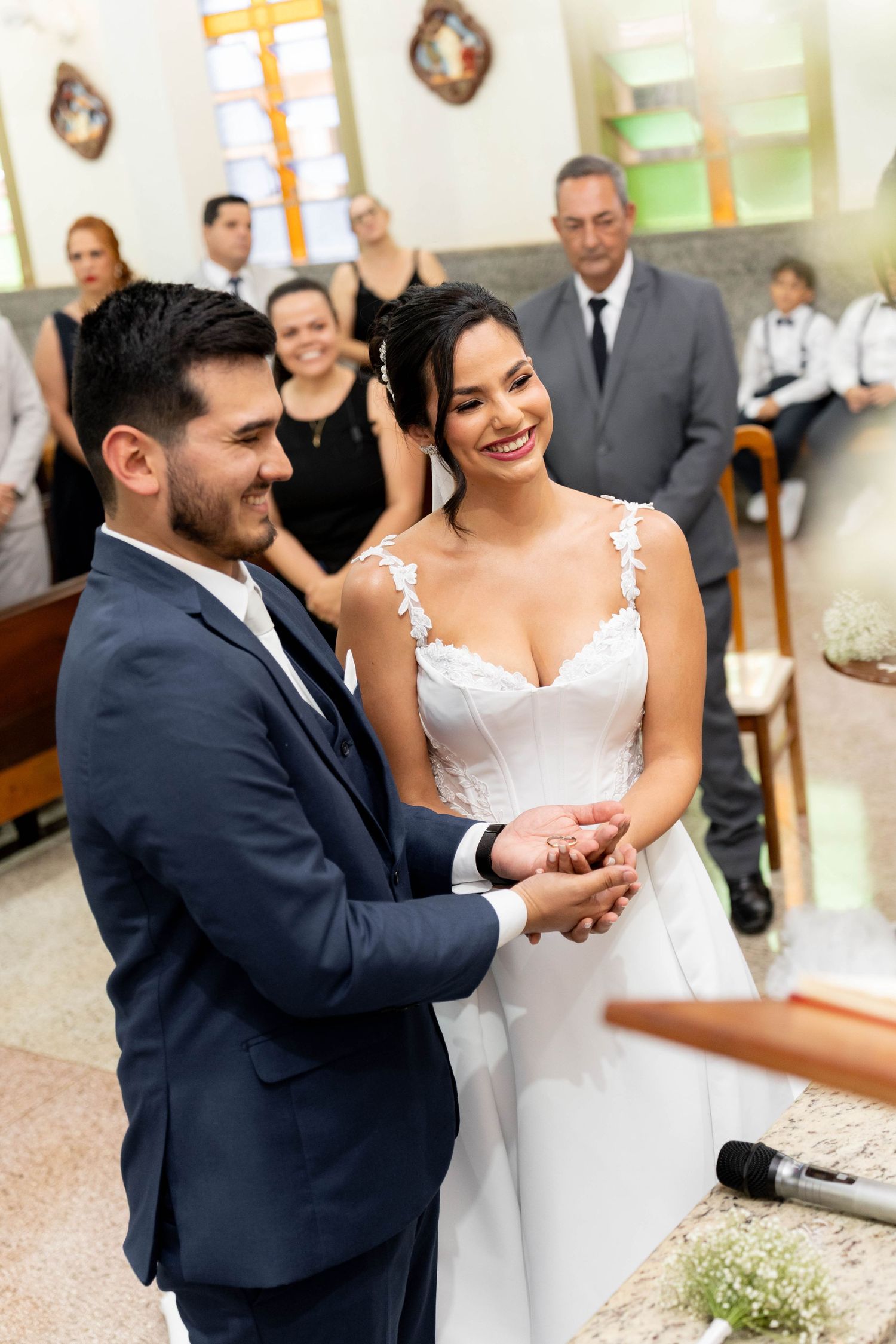 A smiling bride and groom exchange rings during a church wedding ceremony with guests seated in the background.