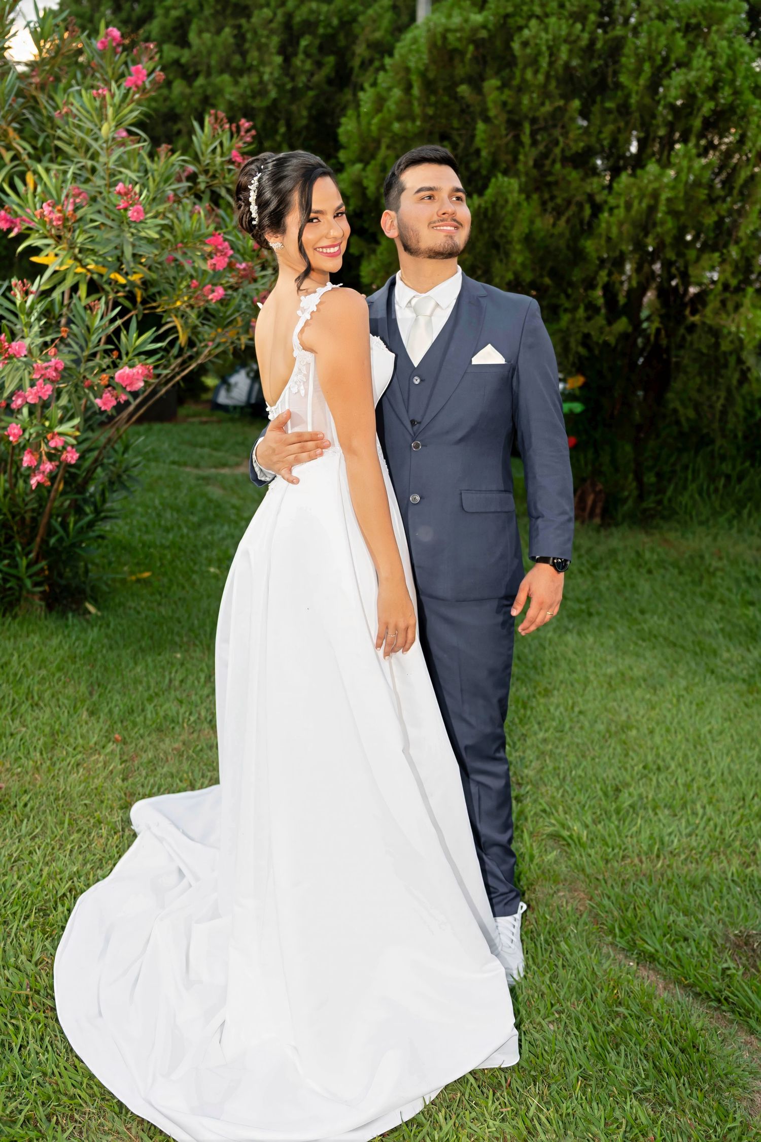 Bride in white gown and groom in gray suit pose together in a lush garden with pink flowers.