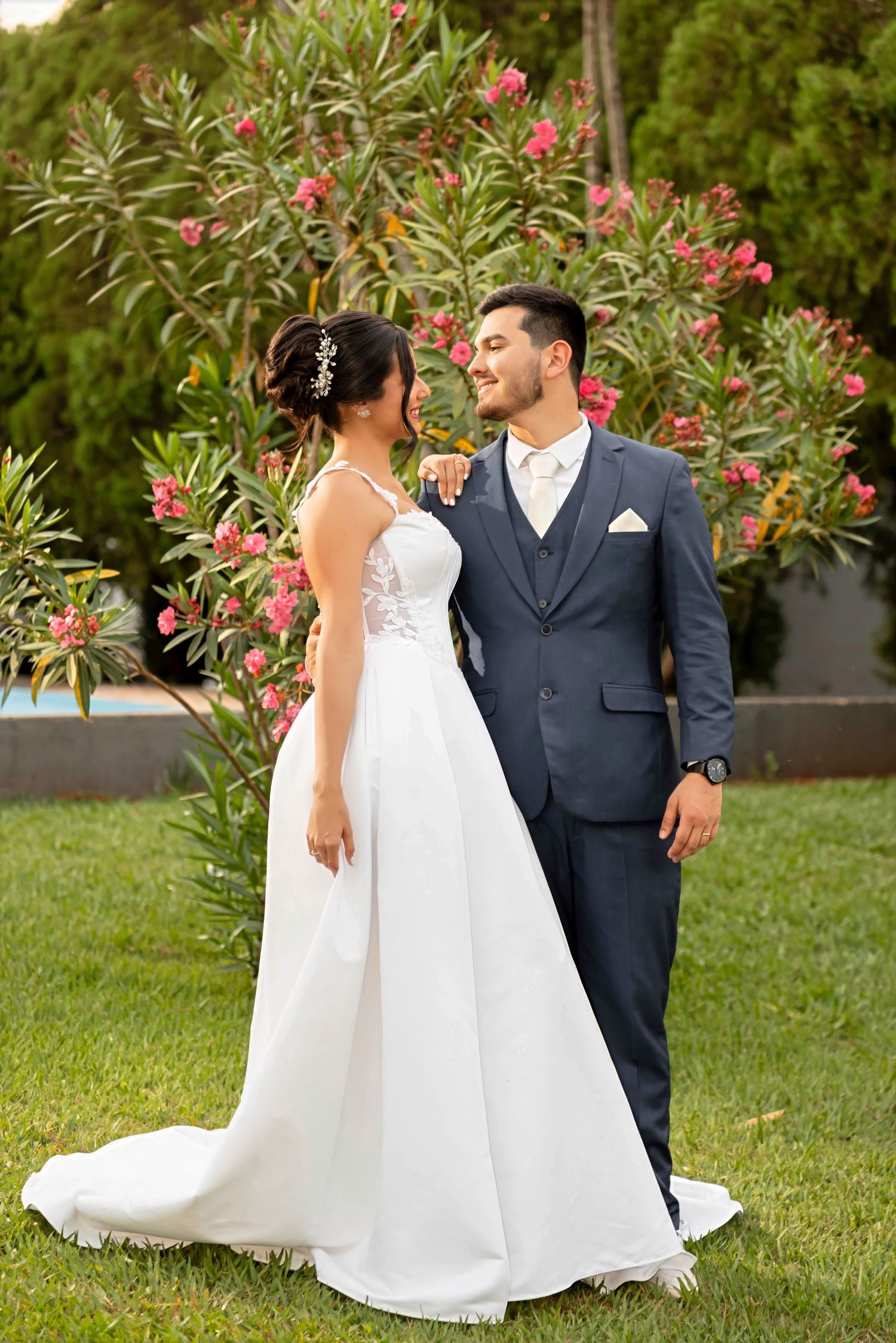 Bride in white gown and groom in navy suit share a romantic moment outdoors near blooming pink flowers.