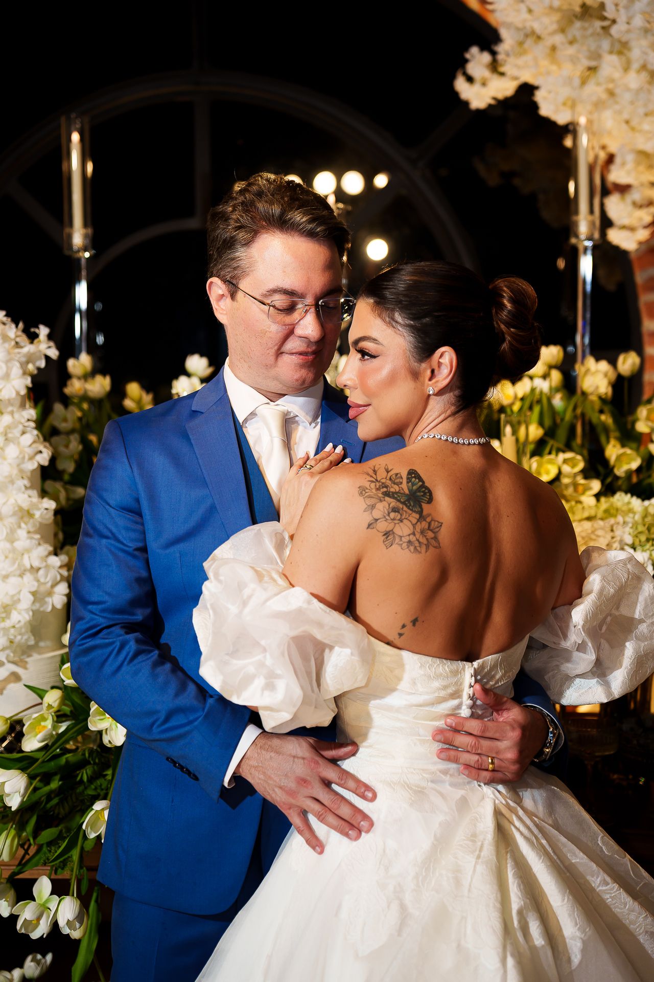 A bride in a backless white gown with tattoos embraces a groom in a blue suit amid white floral wedding decor.