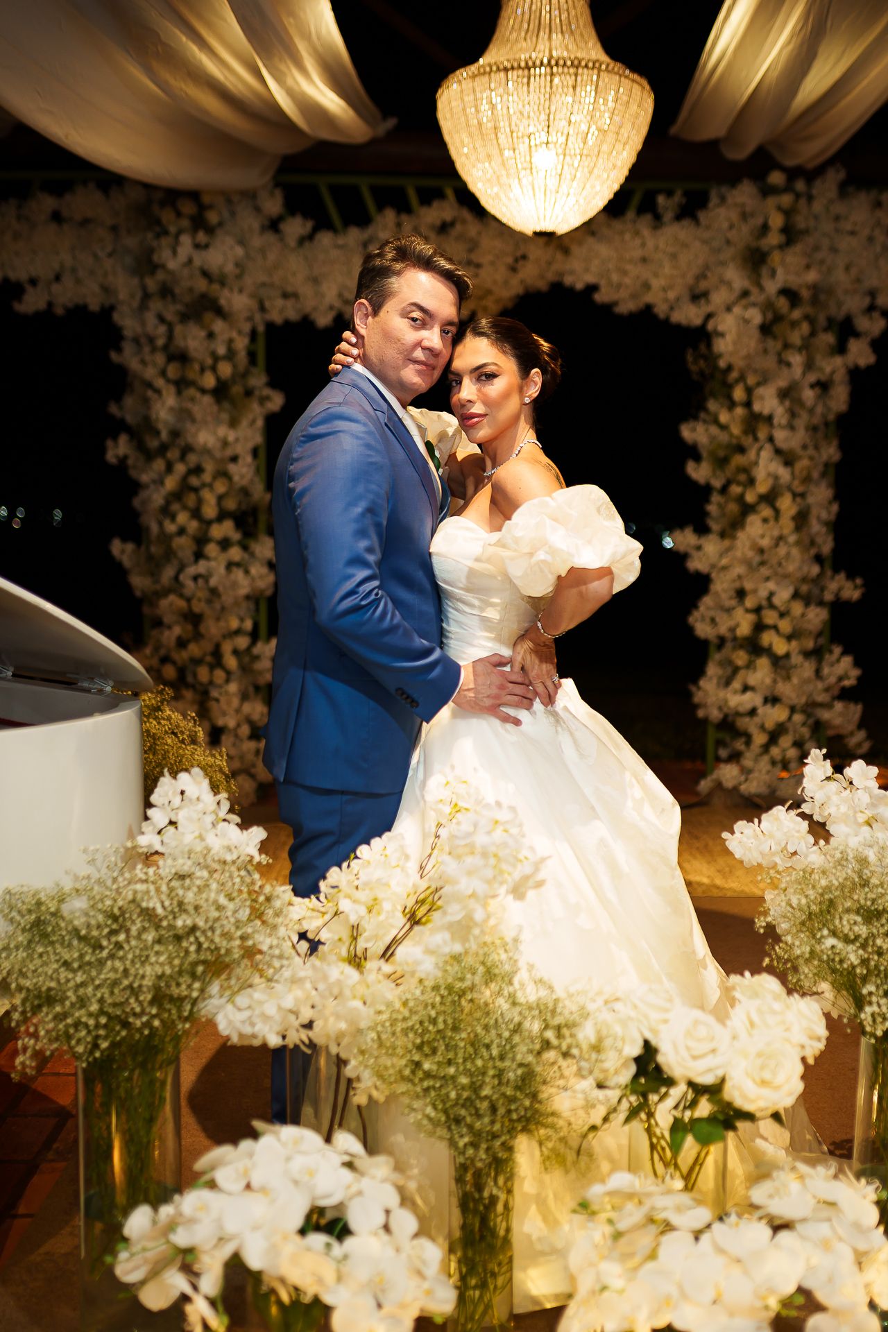 Elegant bride and groom pose together among white floral arrangements under a chandelier at a romantic wedding reception.