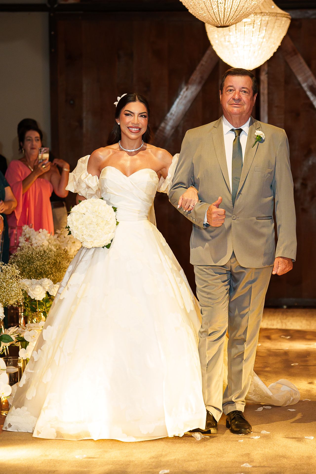 Bride in white ball gown holding bouquet walks down aisle with older man in tan suit at indoor wedding ceremony.