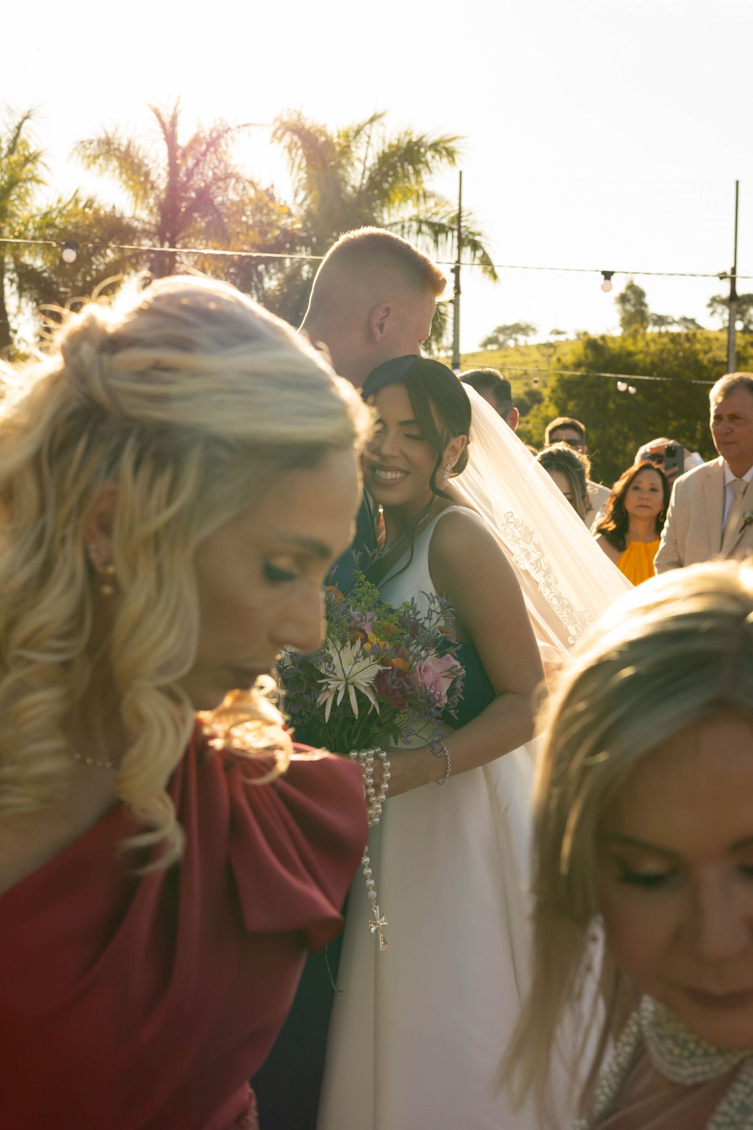 Smiling bride in white gown and veil holds tropical bouquet outdoors at sunny wedding ceremony with guests and palm trees.