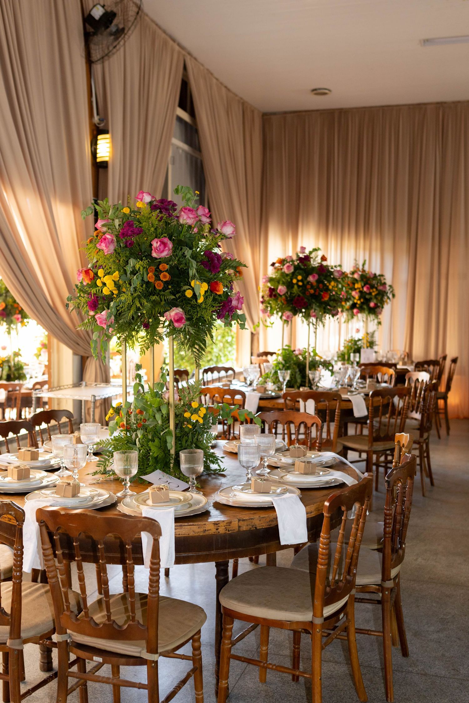 Elegant wedding reception with tall floral centerpieces, wooden chairs, and draped curtains in a warmly lit venue.