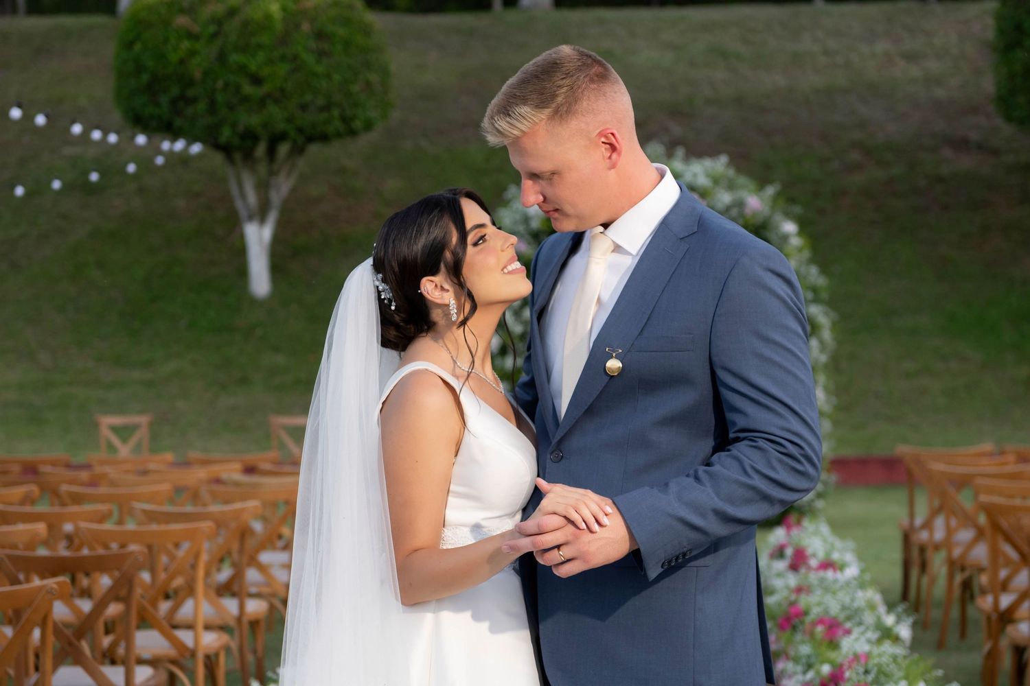 Bride and groom share intimate moment outdoors at wedding venue with wooden chairs and lush green landscape behind them.
