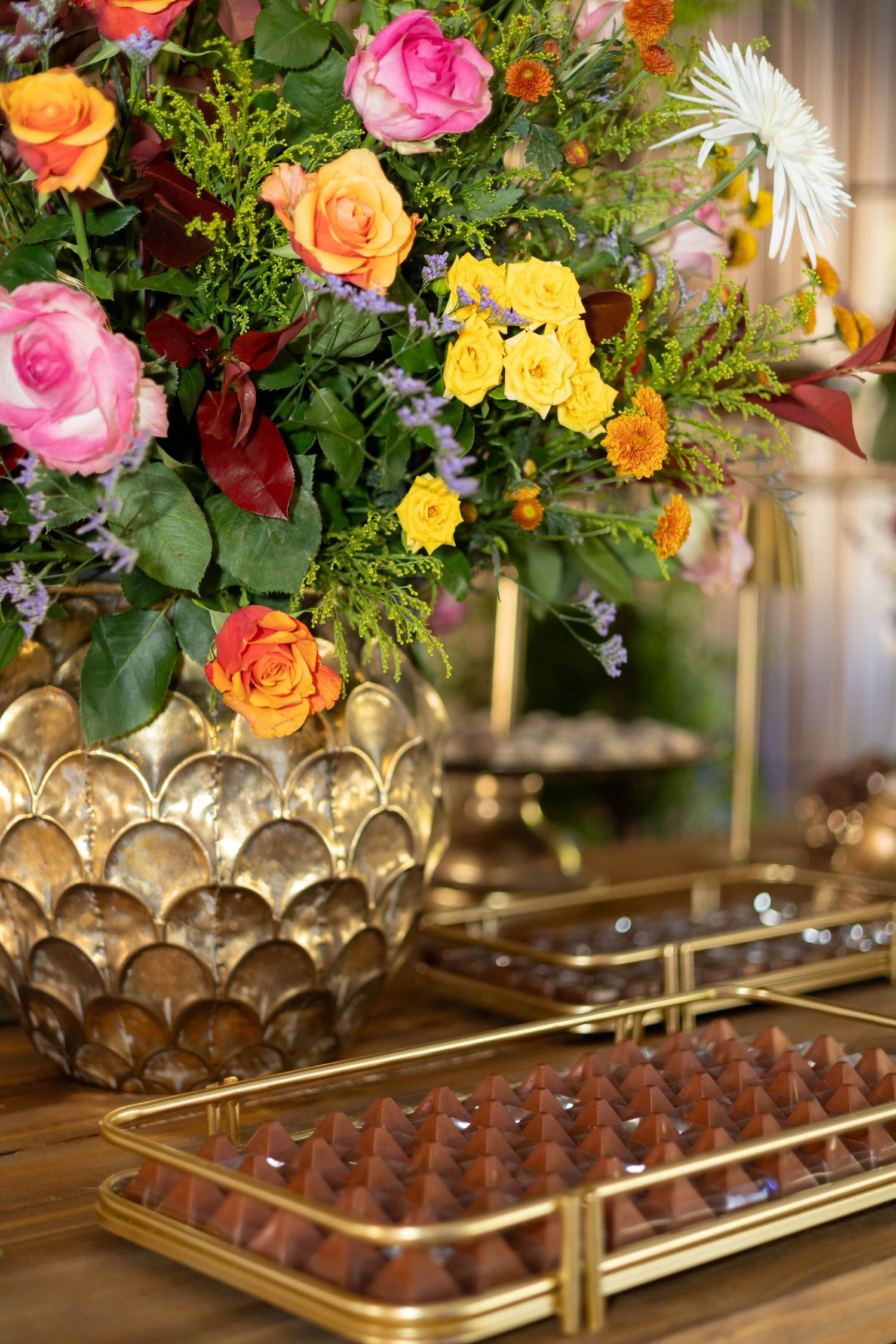 Elegant floral arrangement with colorful roses in gold vase beside trays of chocolates on a wooden table.