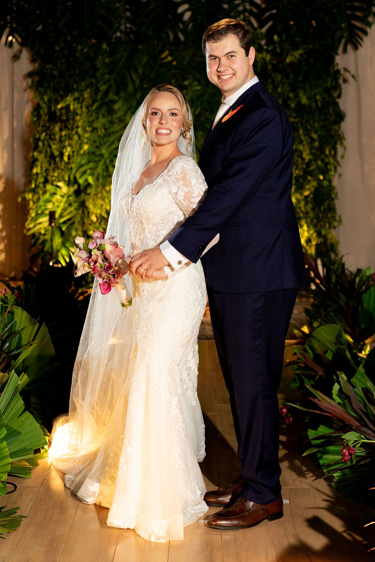 Bride in lace gown with veil holding bouquet stands with groom in navy suit amid tropical greenery at wedding.