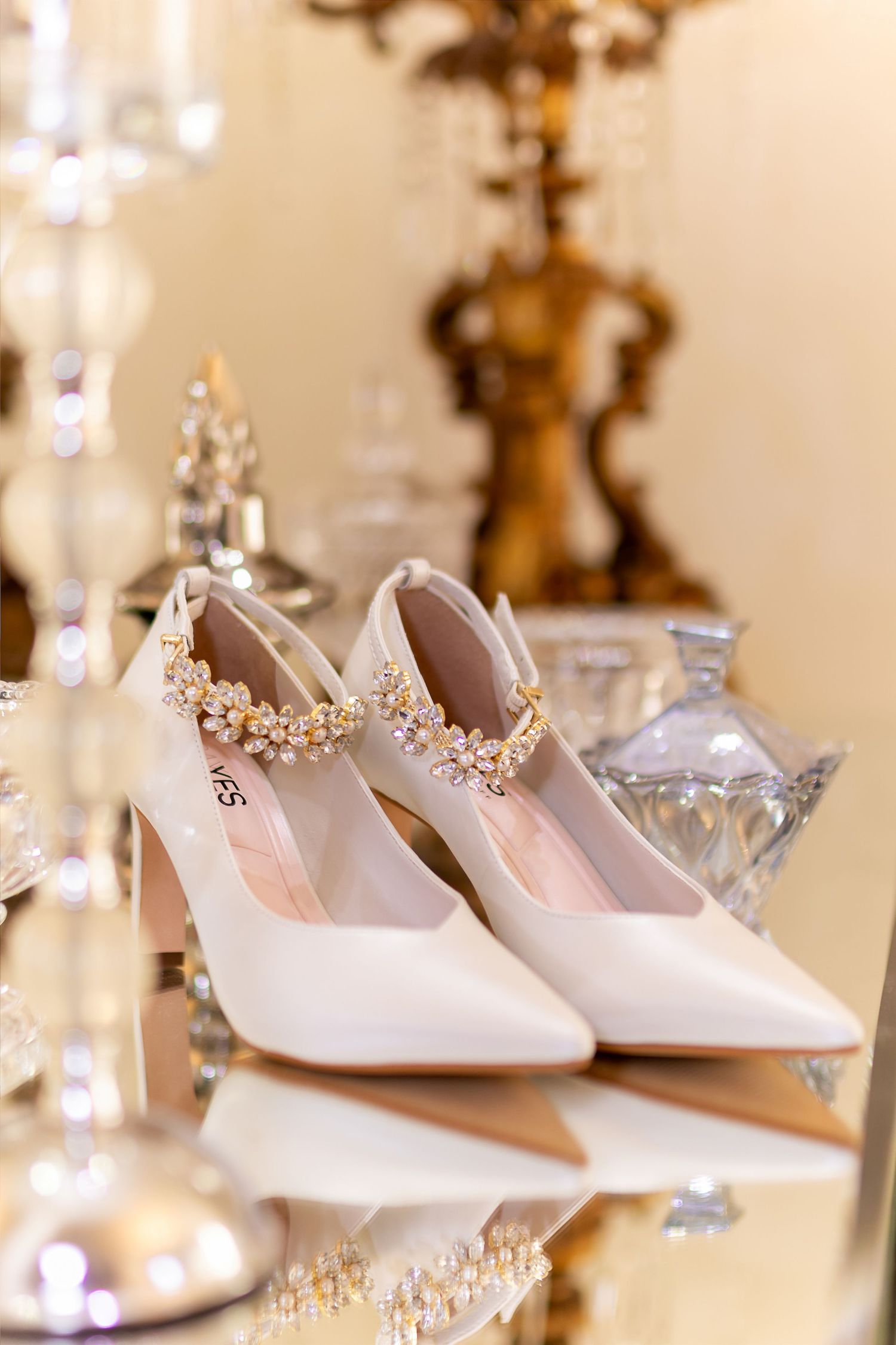 Elegant white bridal heels with gold floral embellishments on a mirrored surface with crystal decor.