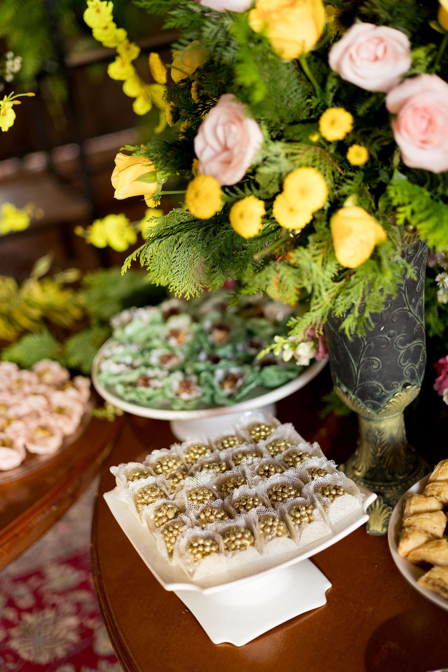 Elegant dessert table with gold-topped chocolates, pink roses, yellow flowers, and greenery in a decorative vase at a wedding reception.