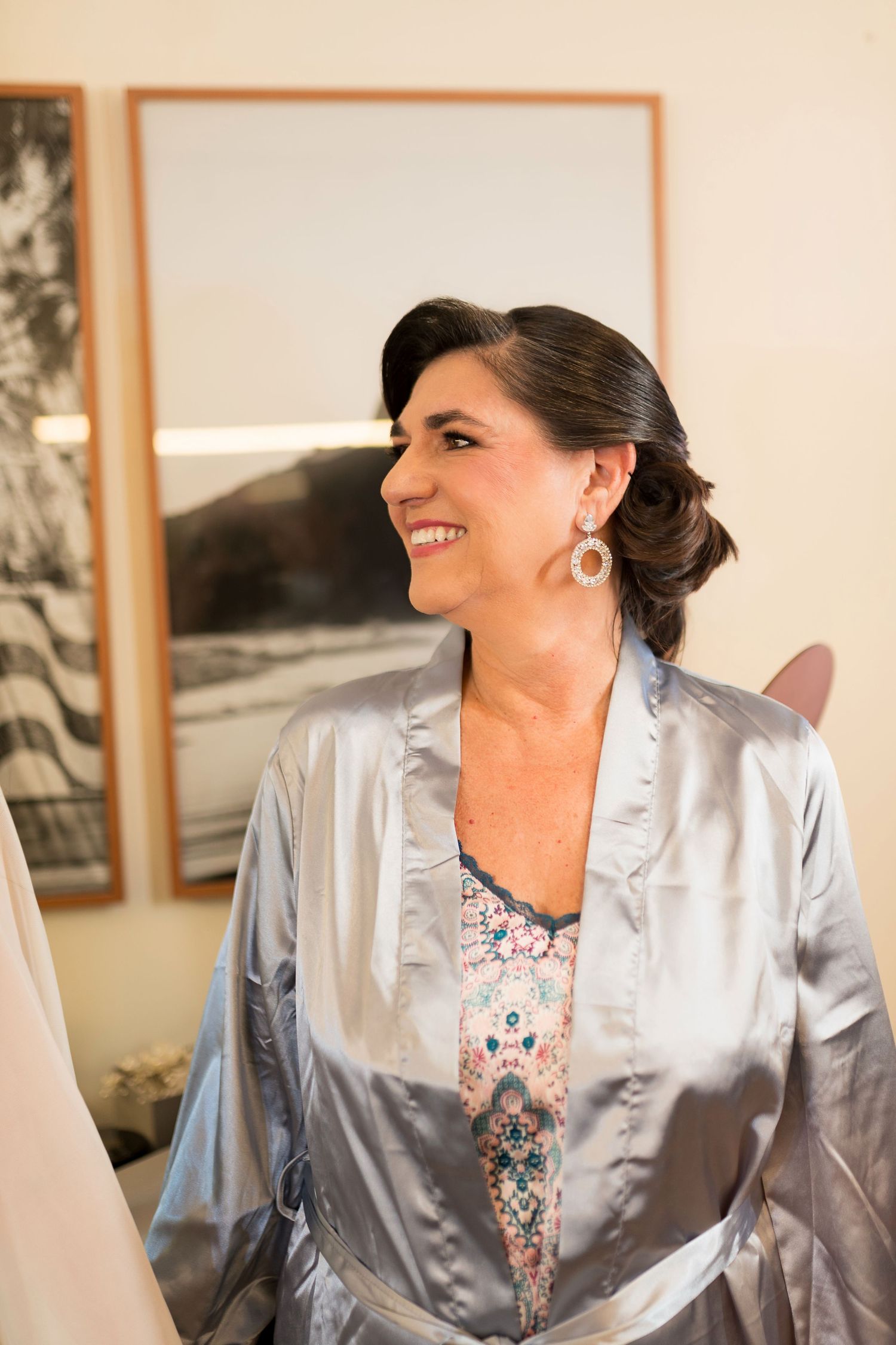 Smiling woman in silver satin robe with floral top and statement earrings, captured in a bright room during bridal preparations.