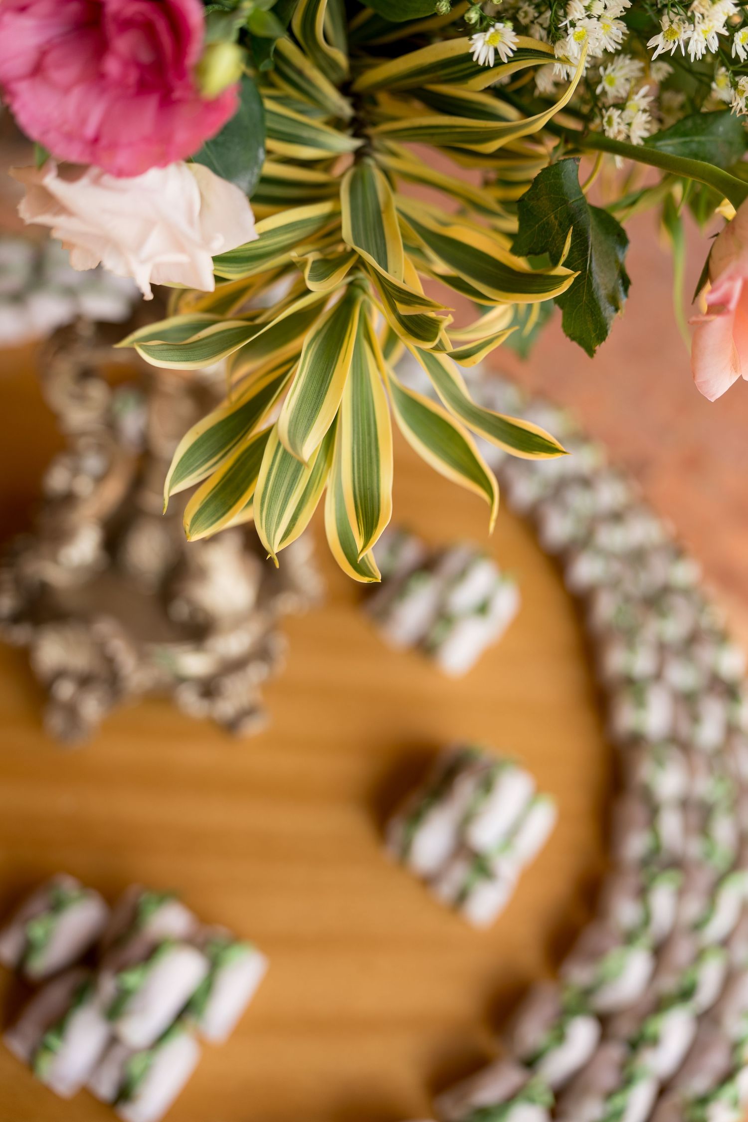 Elegant floral arrangement with pink peonies and tropical leaves above a wooden tray of white chocolate-drizzled treats.