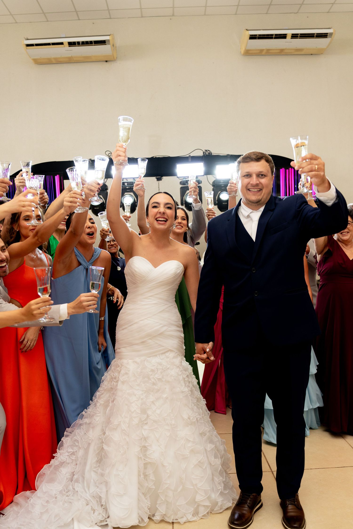 Bride and groom raising champagne glasses with guests lining the aisle at elegant indoor wedding reception.