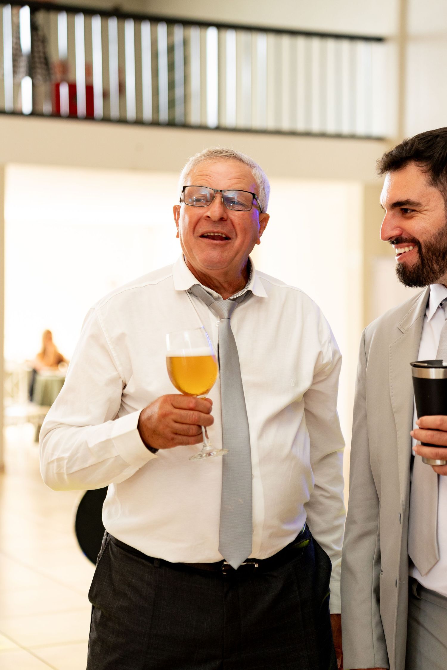 Two men in formal attire smiling and holding drinks at an indoor social gathering.