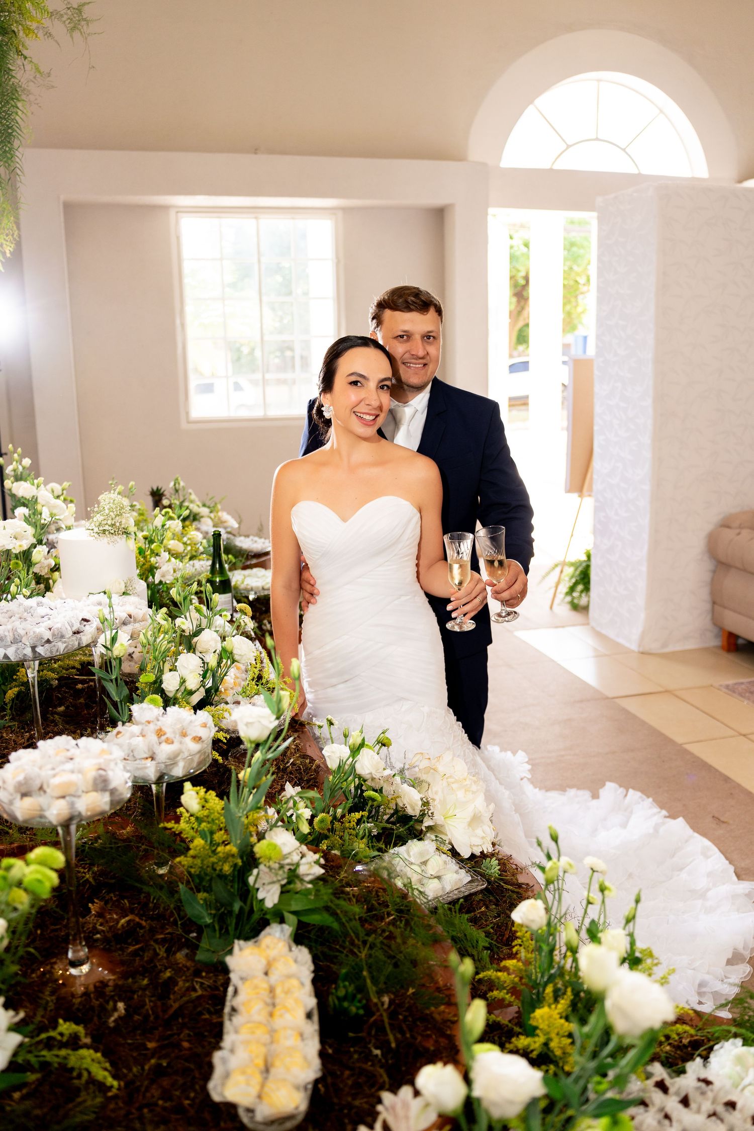 Bride in white gown and groom in navy suit hold champagne glasses among white floral arrangements at their wedding reception.
