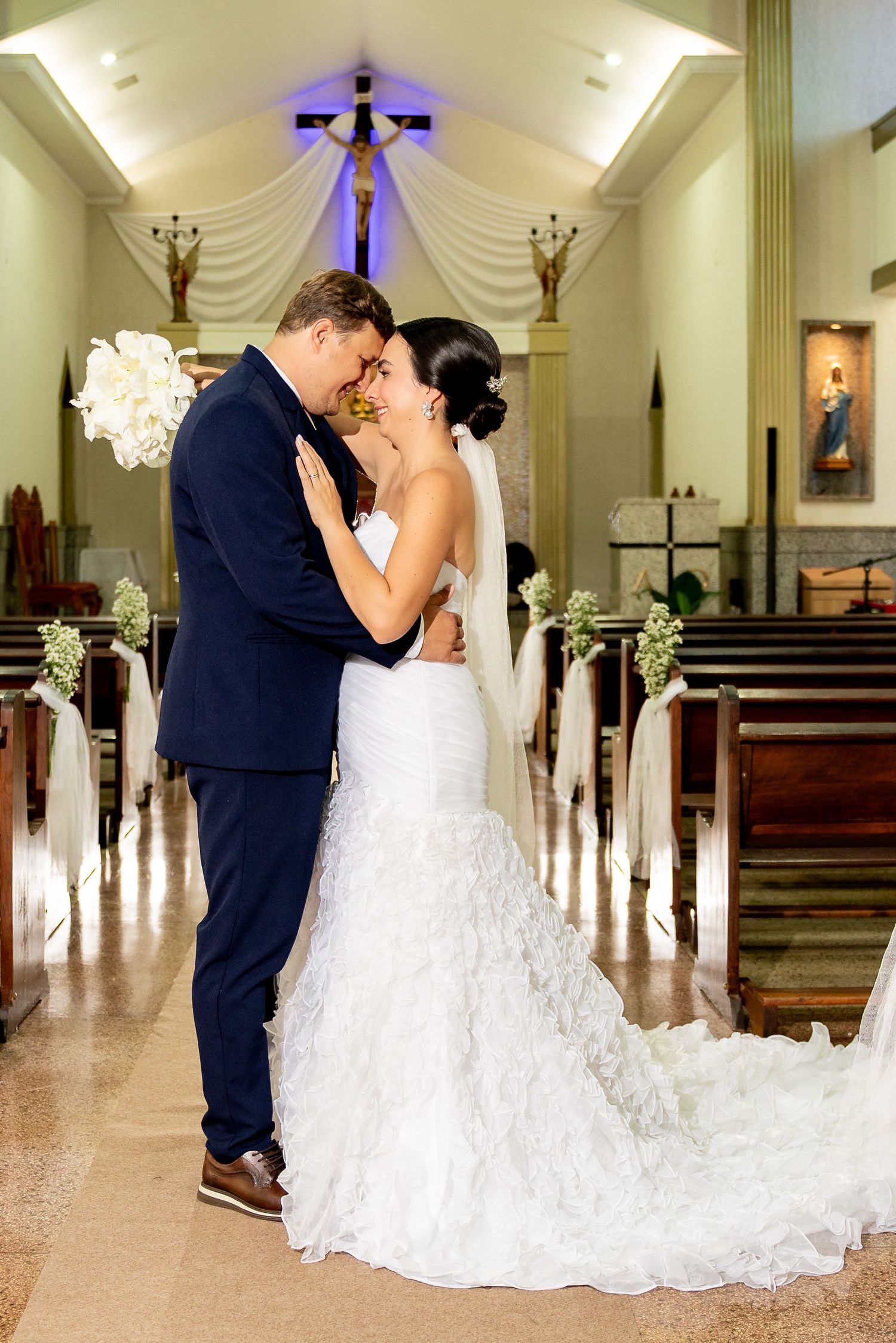 Bride and groom share a romantic embrace in a beautifully decorated church aisle, bride wearing a ruffled mermaid gown.