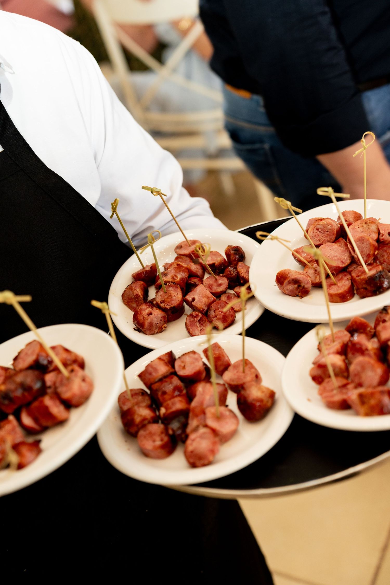 Catered event appetizers of grilled sausage bites on toothpicks served on small white plates by a waiter.