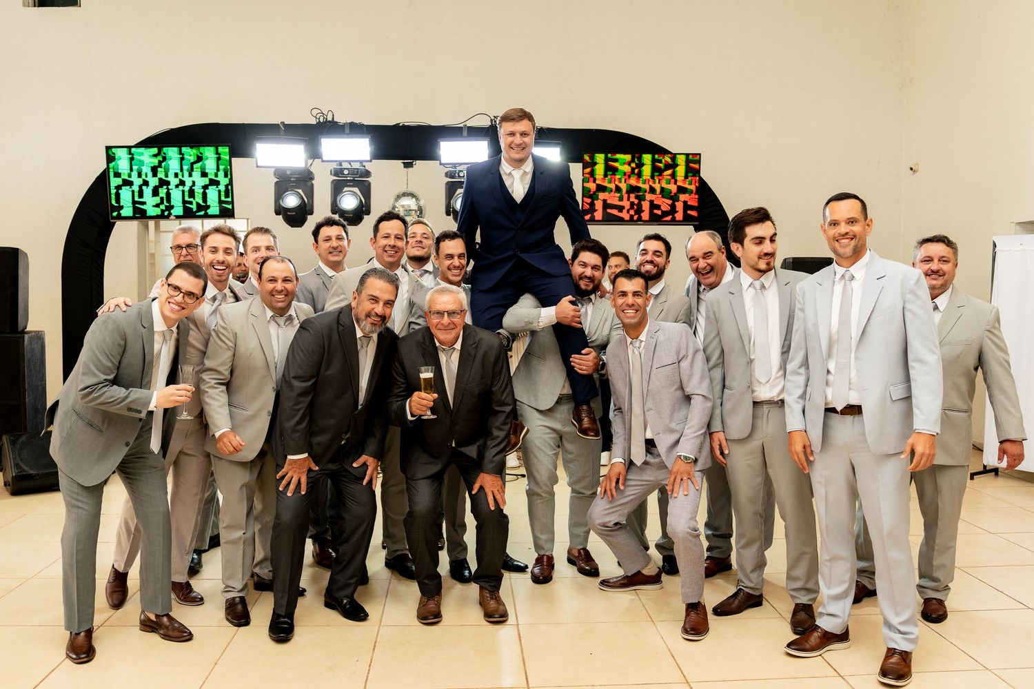 Group of men in gray suits celebrating at a wedding reception, lifting the groom in the air while smiling.