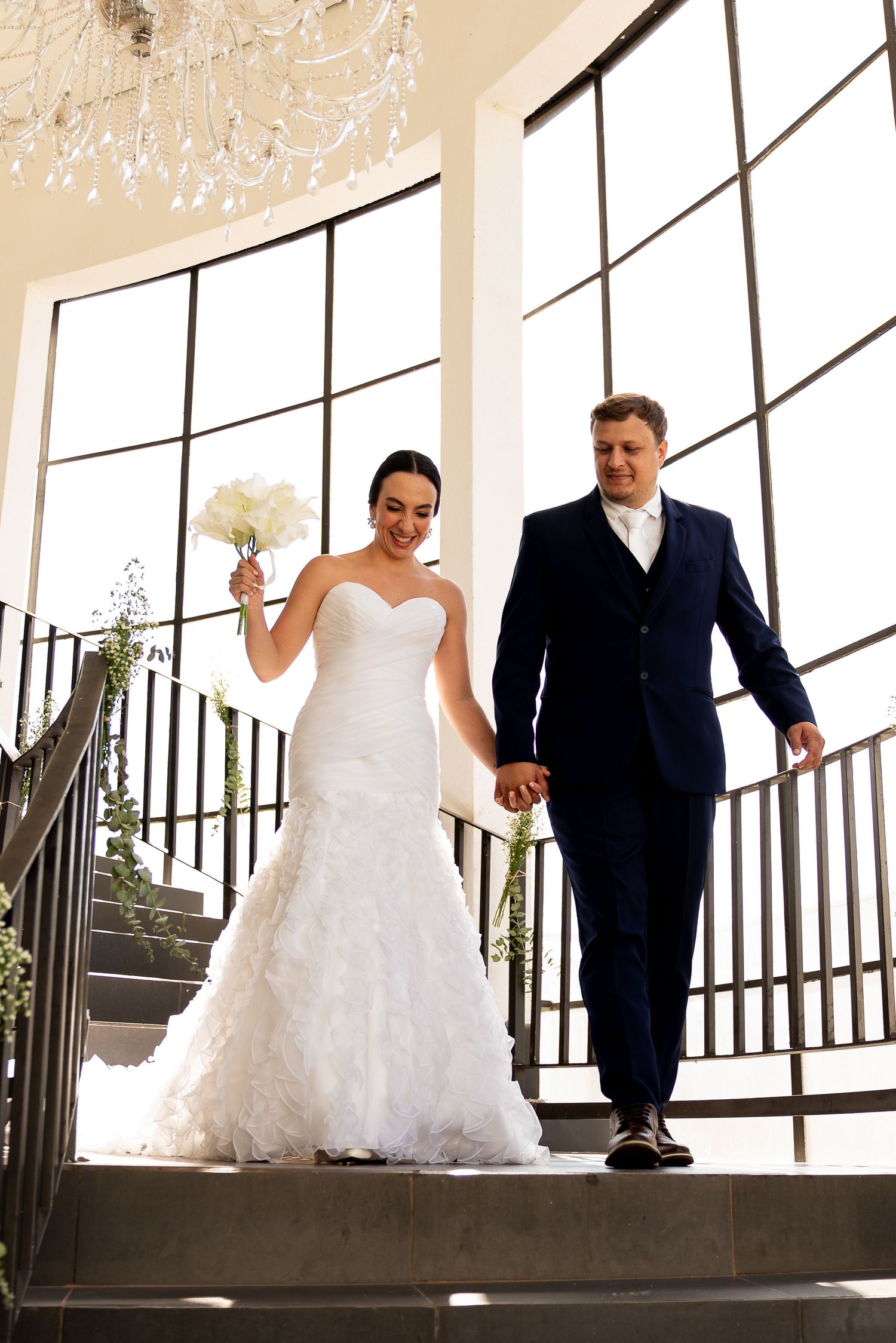 Bride in white mermaid gown and groom in navy suit descend elegant staircase holding hands at wedding venue.