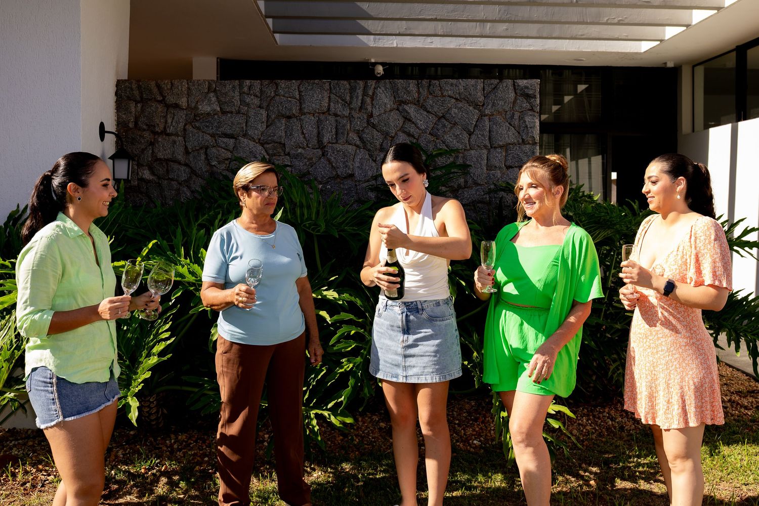 Five women socializing outdoors, holding drinks and smiling near tropical plants and a stone wall.