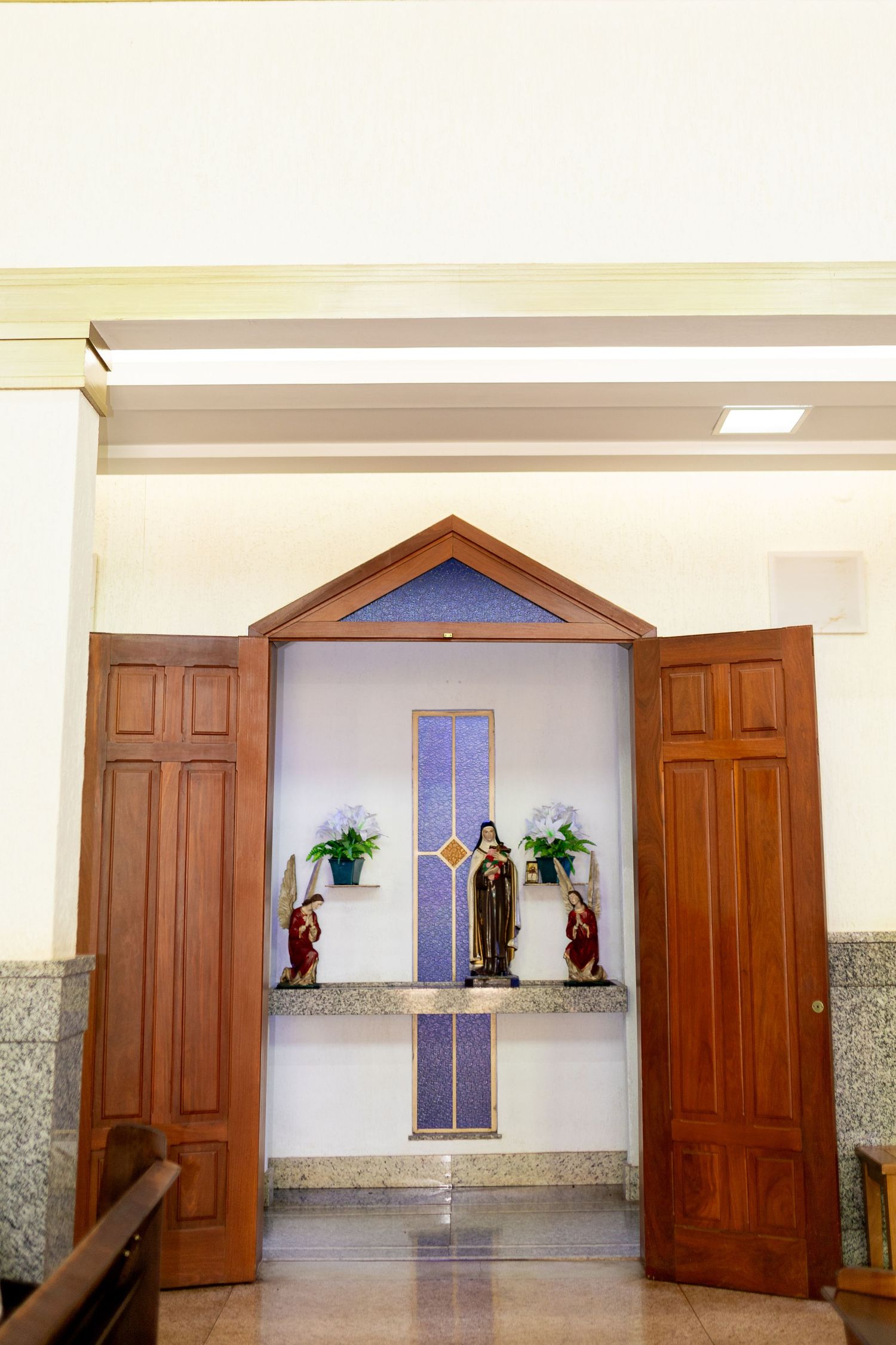 Church interior with open wooden doors revealing a small altar with religious statue and floral arrangements.