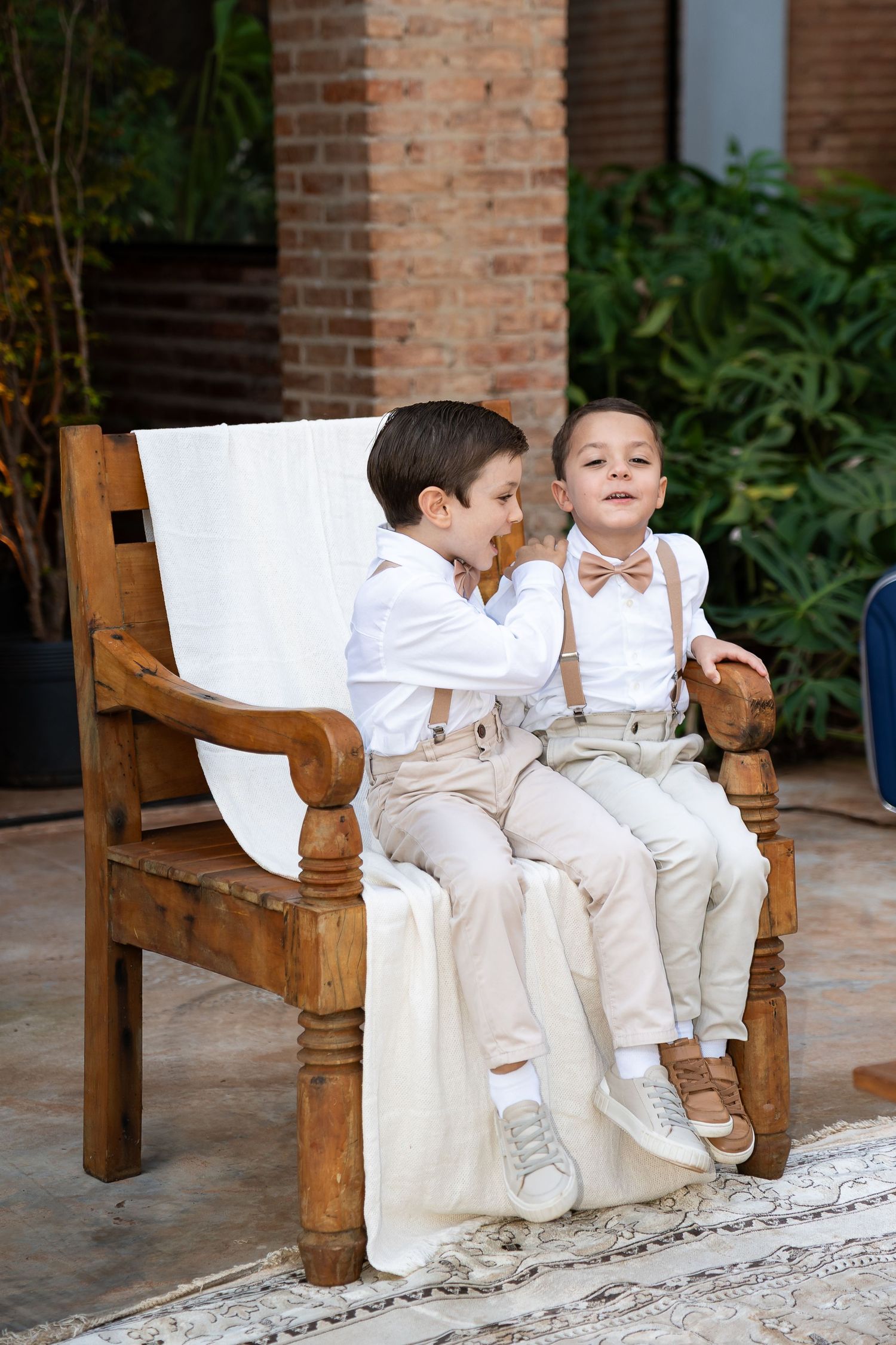 Two young boys in matching white shirts, suspenders, and bow ties sitting on a wooden bench at a formal event.