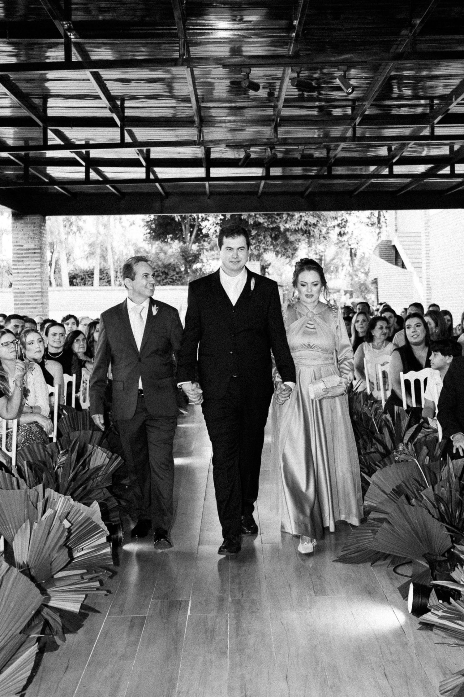 Groom walks down wedding aisle escorted by parents in black and white ceremony photo.