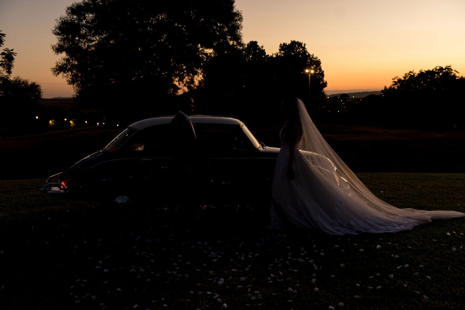Silhouette of bride in flowing veil leaning against car at sunset during golden hour outdoor wedding.
