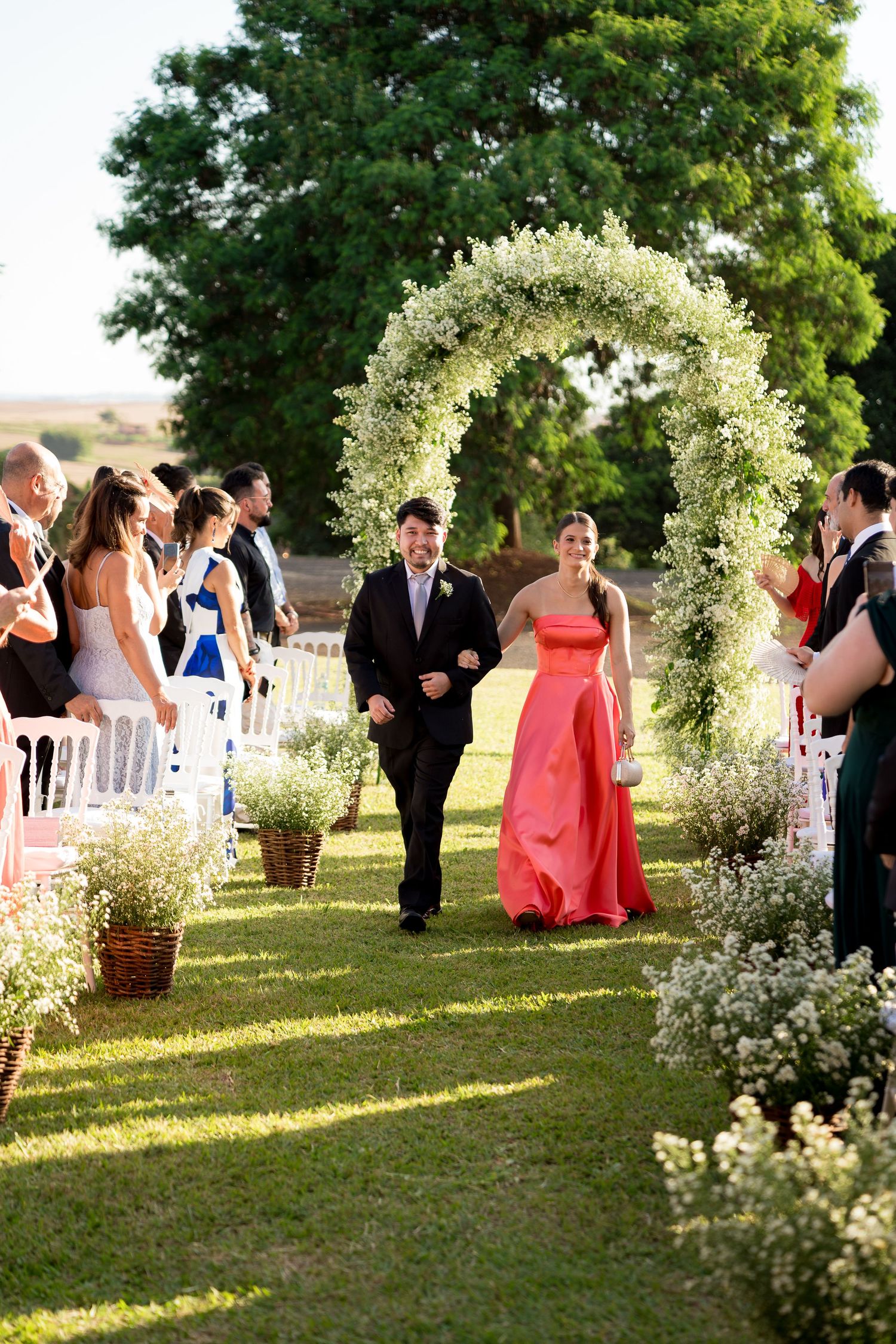 Couple walks down outdoor wedding aisle under floral arch, guests applaud, surrounded by white flowers and lush greenery.