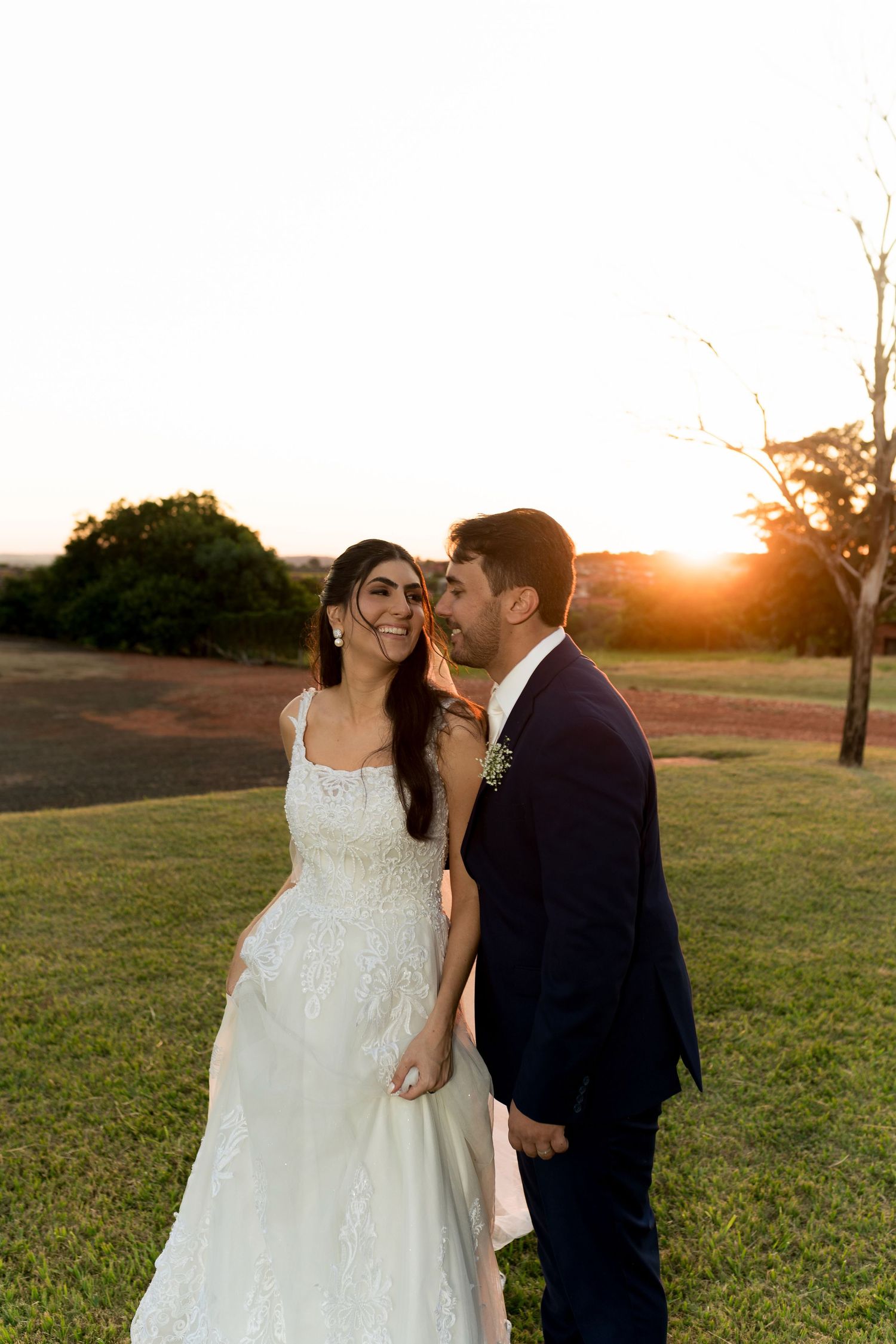 Bride in white lace gown and groom in navy suit share a romantic sunset moment on green lawn.