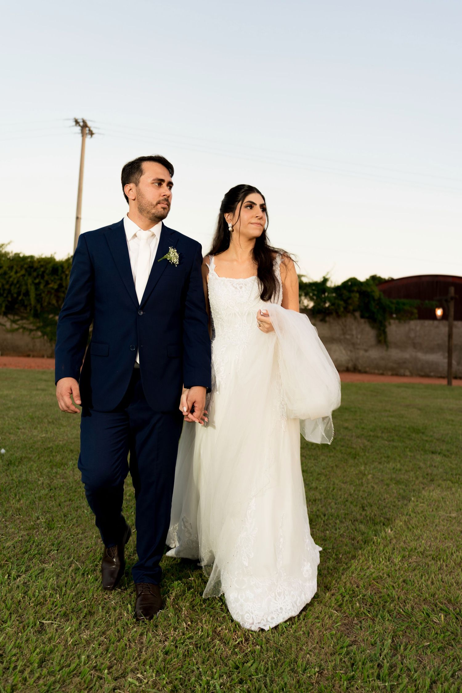 Bride in white gown and groom in navy suit walk hand in hand on green grass at outdoor wedding venue.