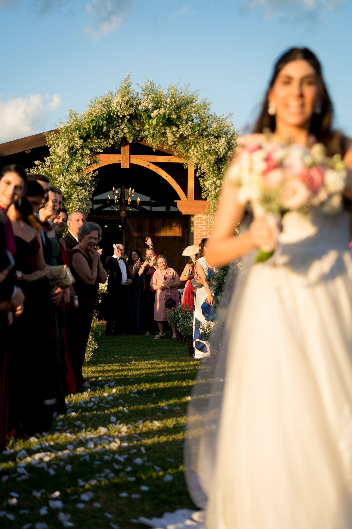 Bride in white gown holding floral bouquet walks down outdoor aisle as guests applaud near floral arch.