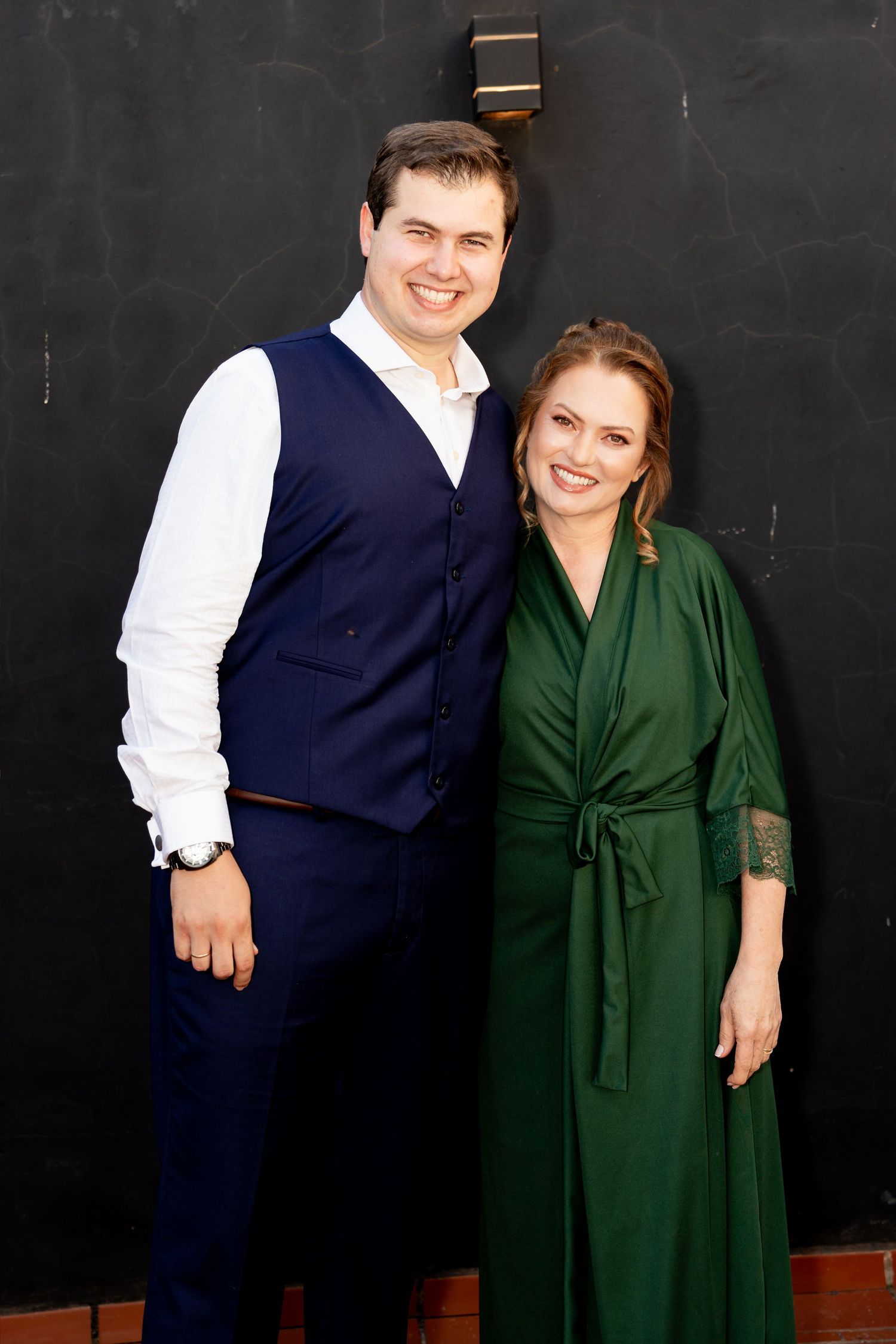 Smiling couple posing together, man in blue vest and white shirt, woman in elegant green wrap dress, against dark wall.