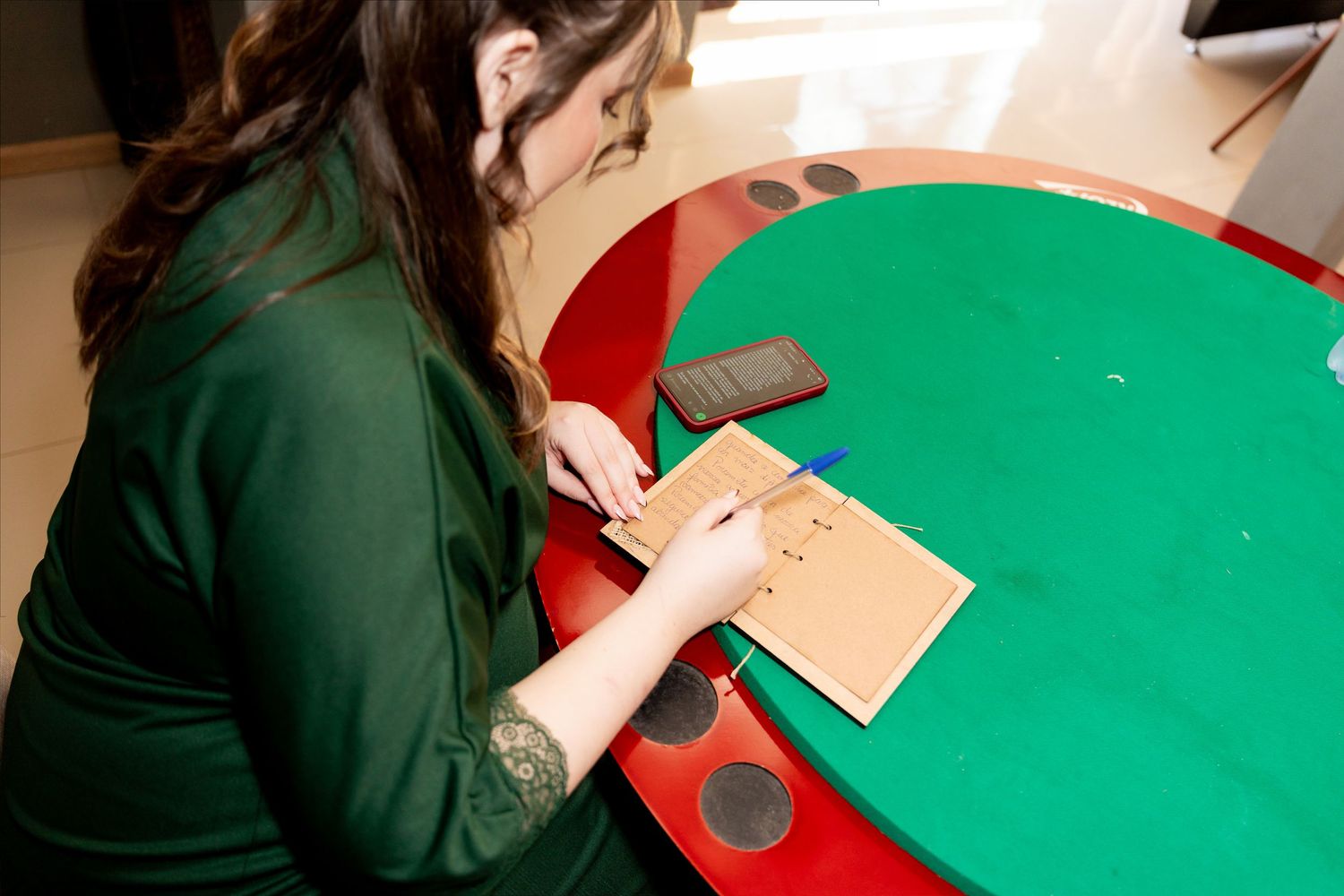 Woman in green dress writing on a brown envelope at a colorful round table.