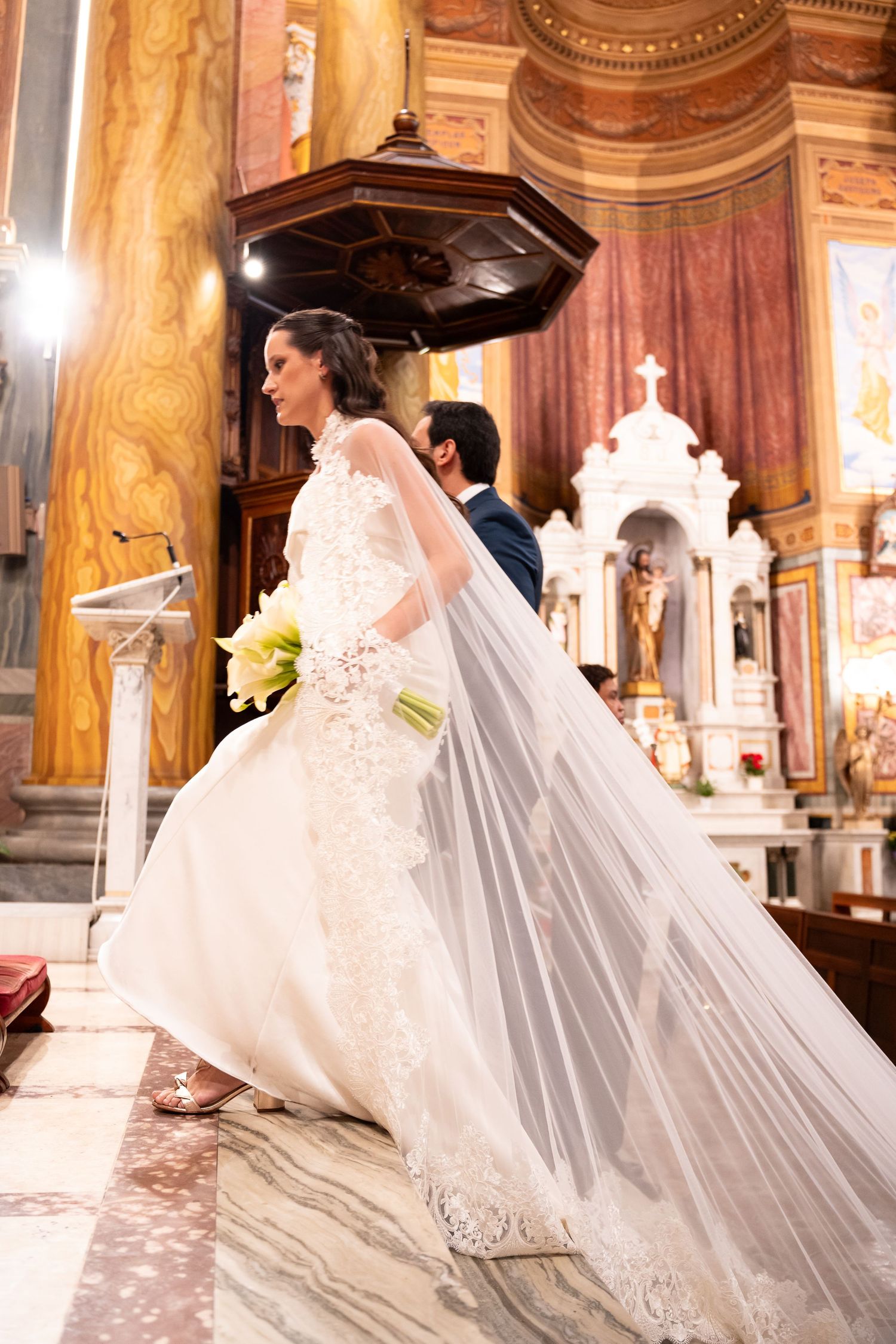Bride in lace wedding gown and long veil walks down church aisle holding yellow bouquet during ceremony.