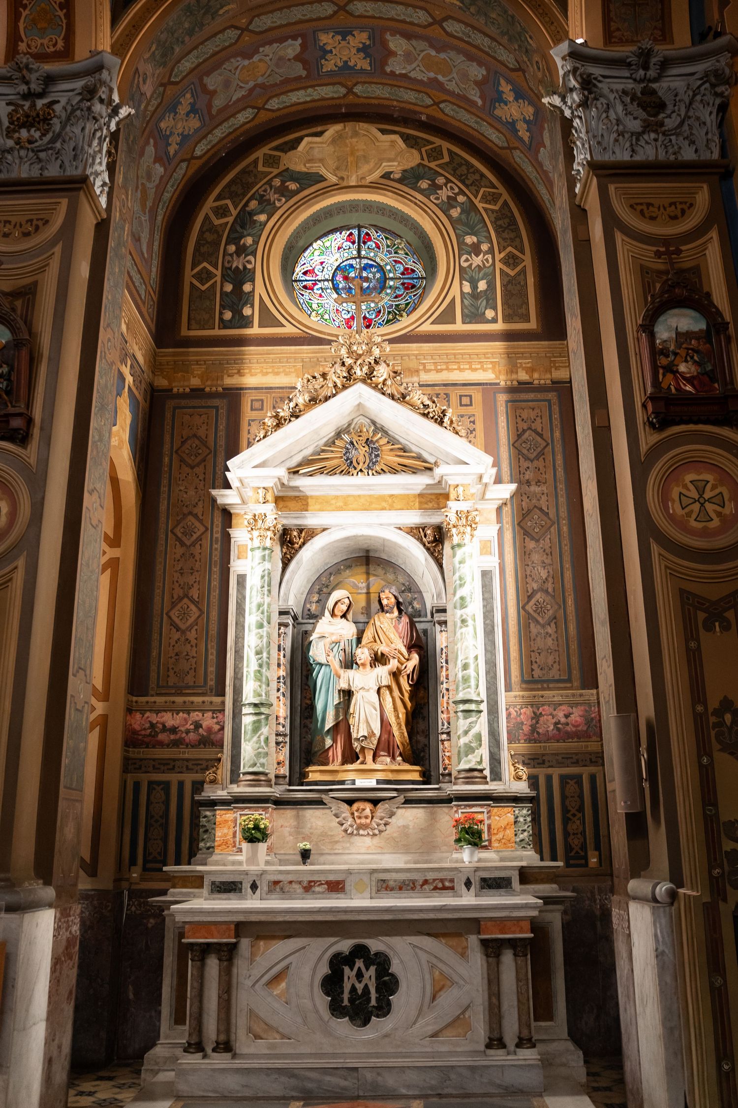 Ornate Catholic church altar with marble statues of Holy Family beneath a decorative arch and stained glass rose window.