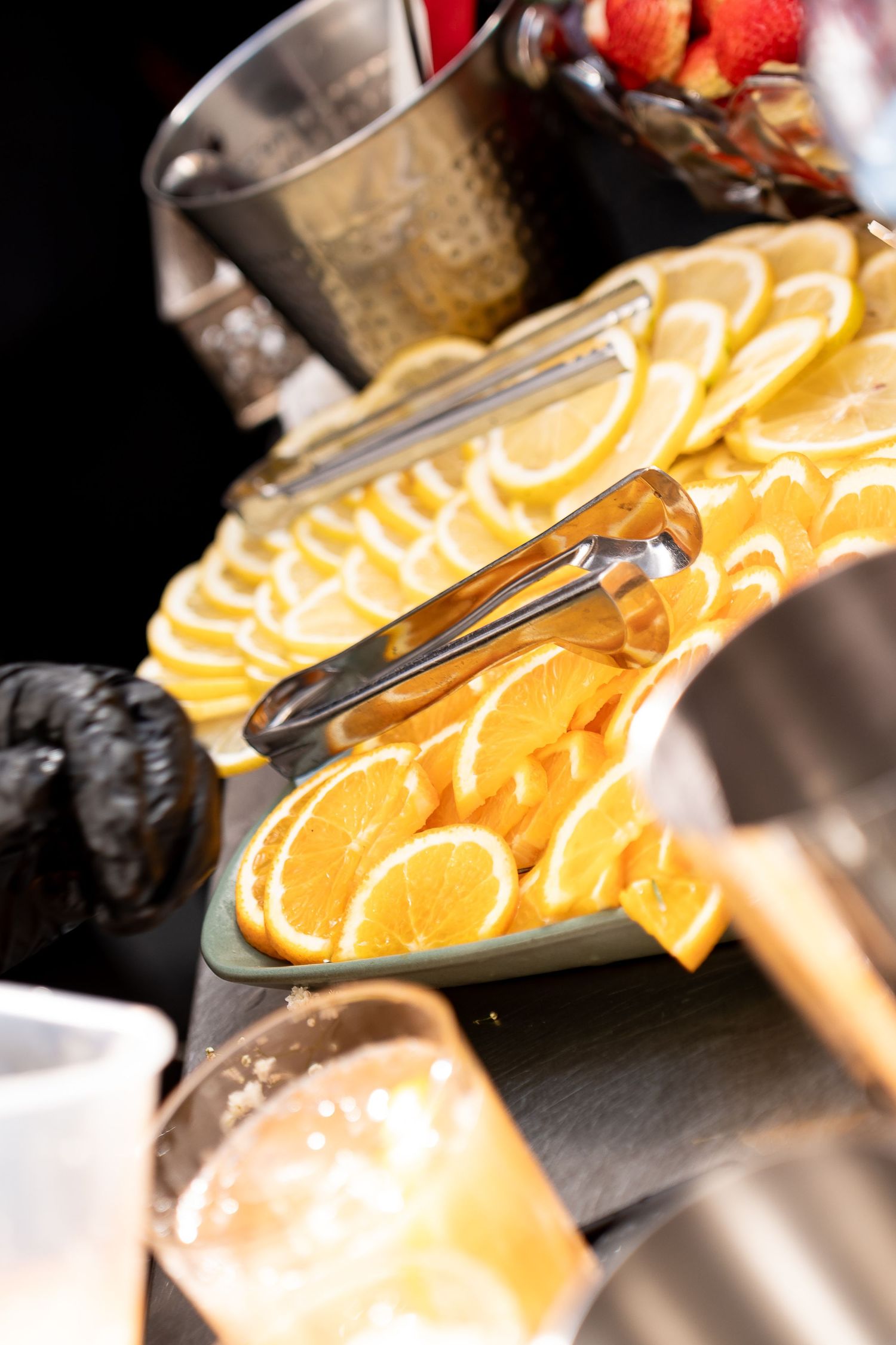 Sliced oranges and lemons arranged on a platter with tongs at a beverage station, surrounded by glasses and garnishes.