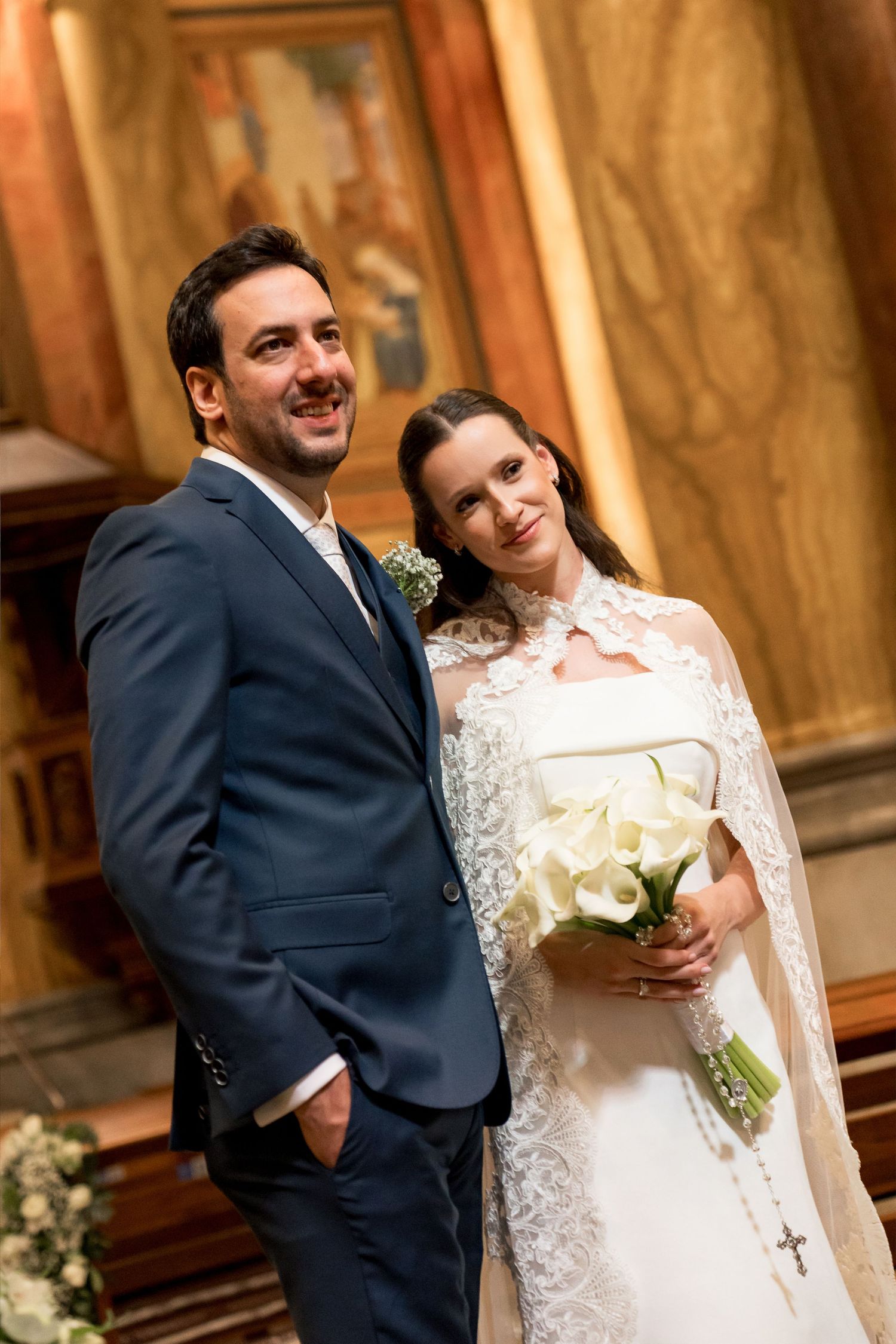 A smiling bride holding white calla lilies bouquet stands beside groom in navy suit inside elegant church.