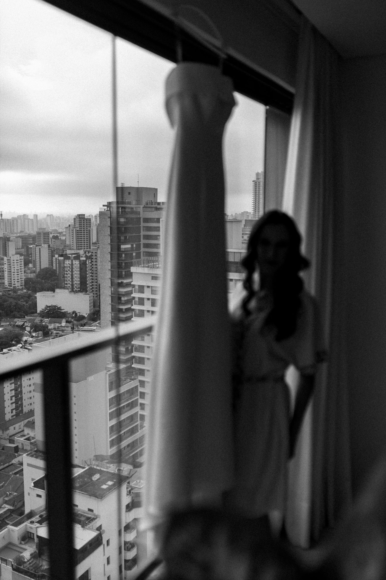 Black and white photo of a bride in robe beside her hanging wedding gown, with urban skyline visible through window.